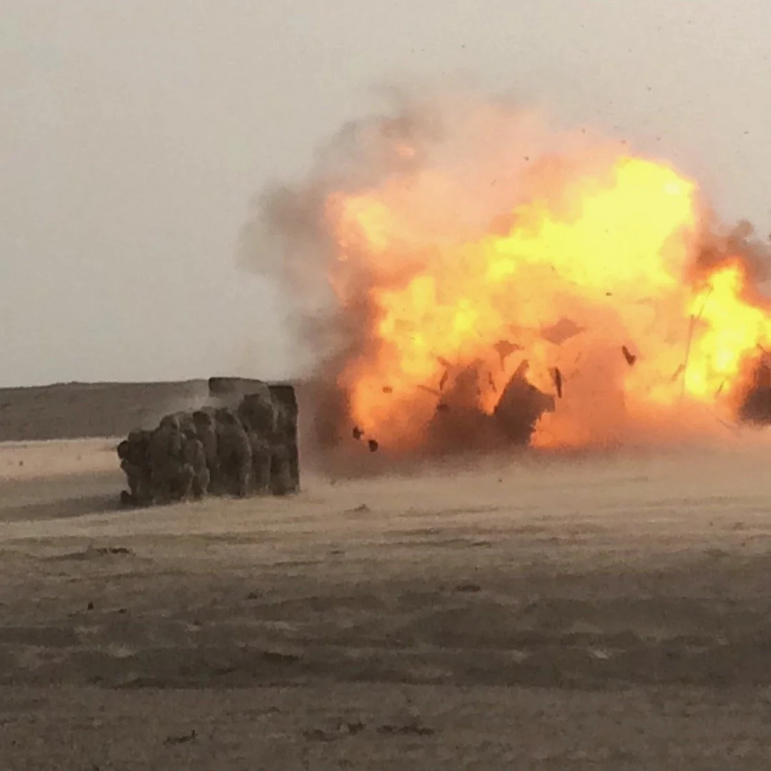 Explosion with fire and smoke in a desert landscape, with a group of Marines near the explosion.