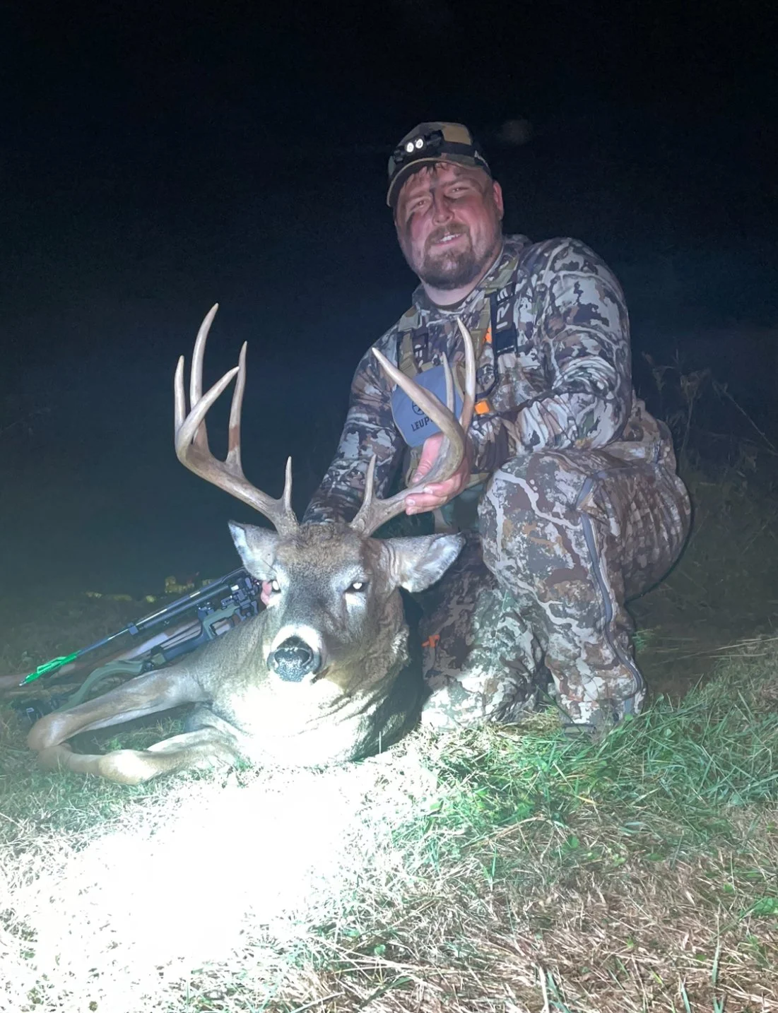 A man dressed in camouflage hunting gear at night kneeling next to a large deer with antlers, holding its antlers while posing for the photo.