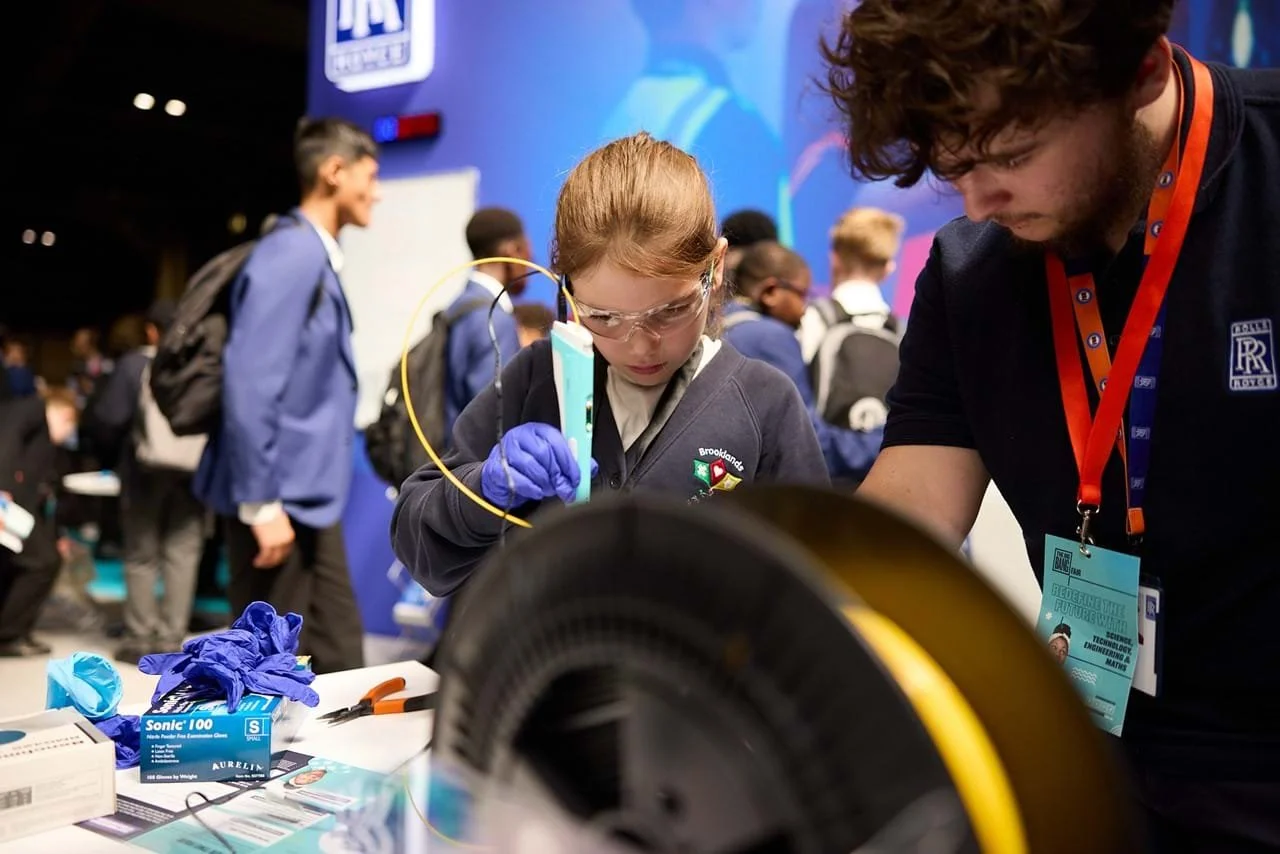A girl with safety goggles and gloves working on a scientific project at a science fair, with a man assisting her, and students in the background.