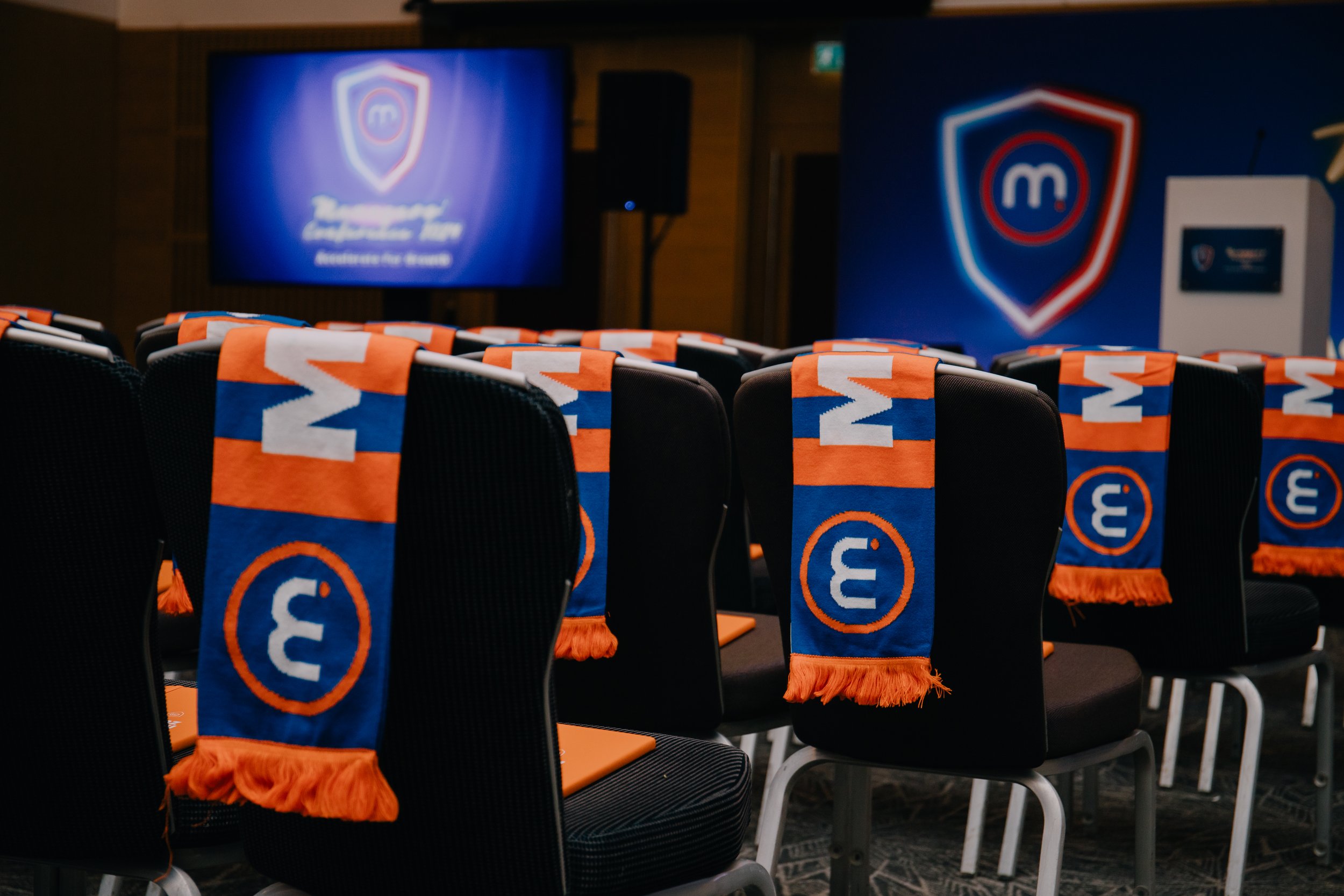 Conference room with chairs draped with orange and blue scarves featuring a logo and the number 3, facing a stage with a large screen displaying a shield logo and text.