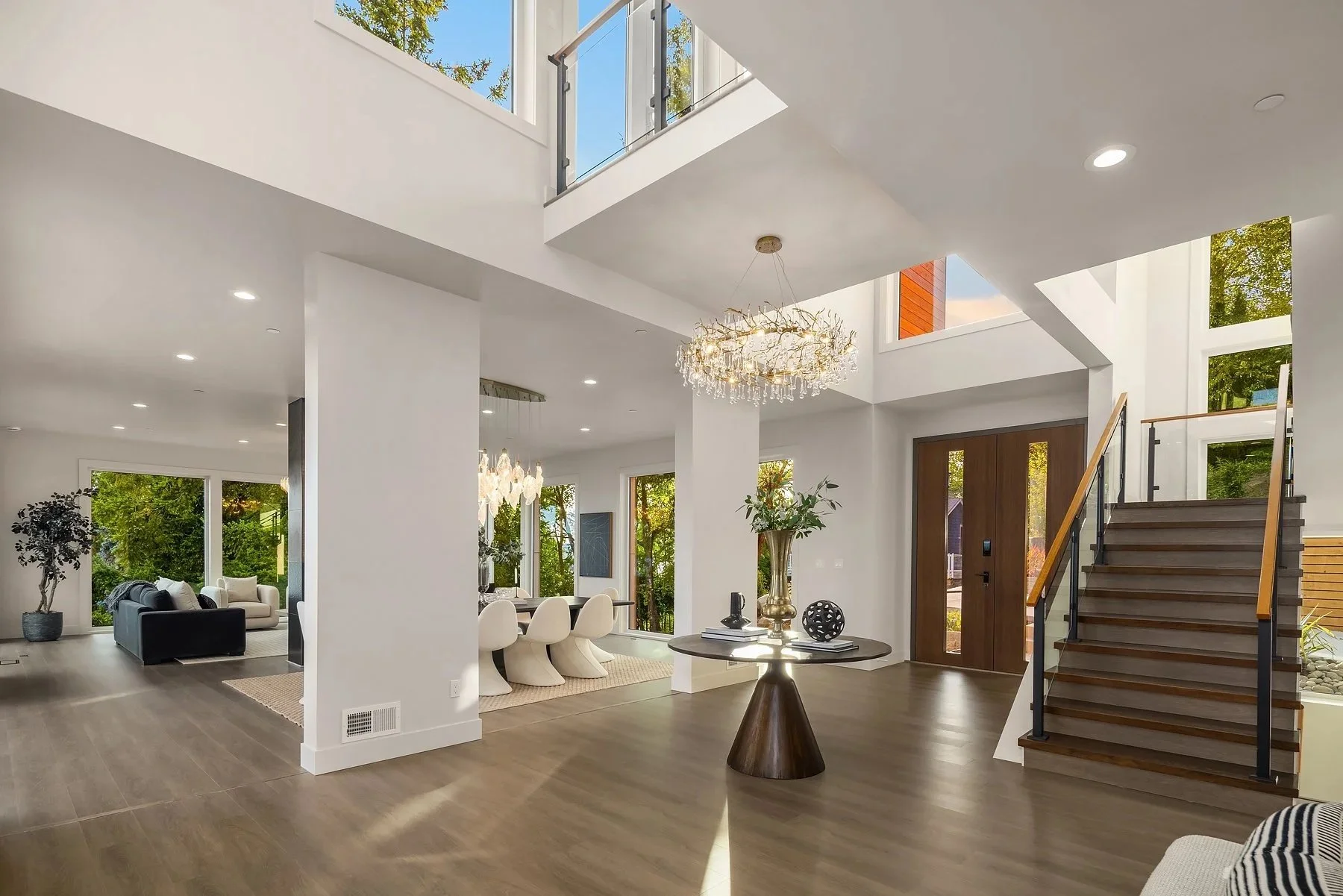 Interior view of a modern, spacious home with open floor plan, featuring a staircase, large windows, a chandelier, and living and dining areas with stylish furniture and decor.