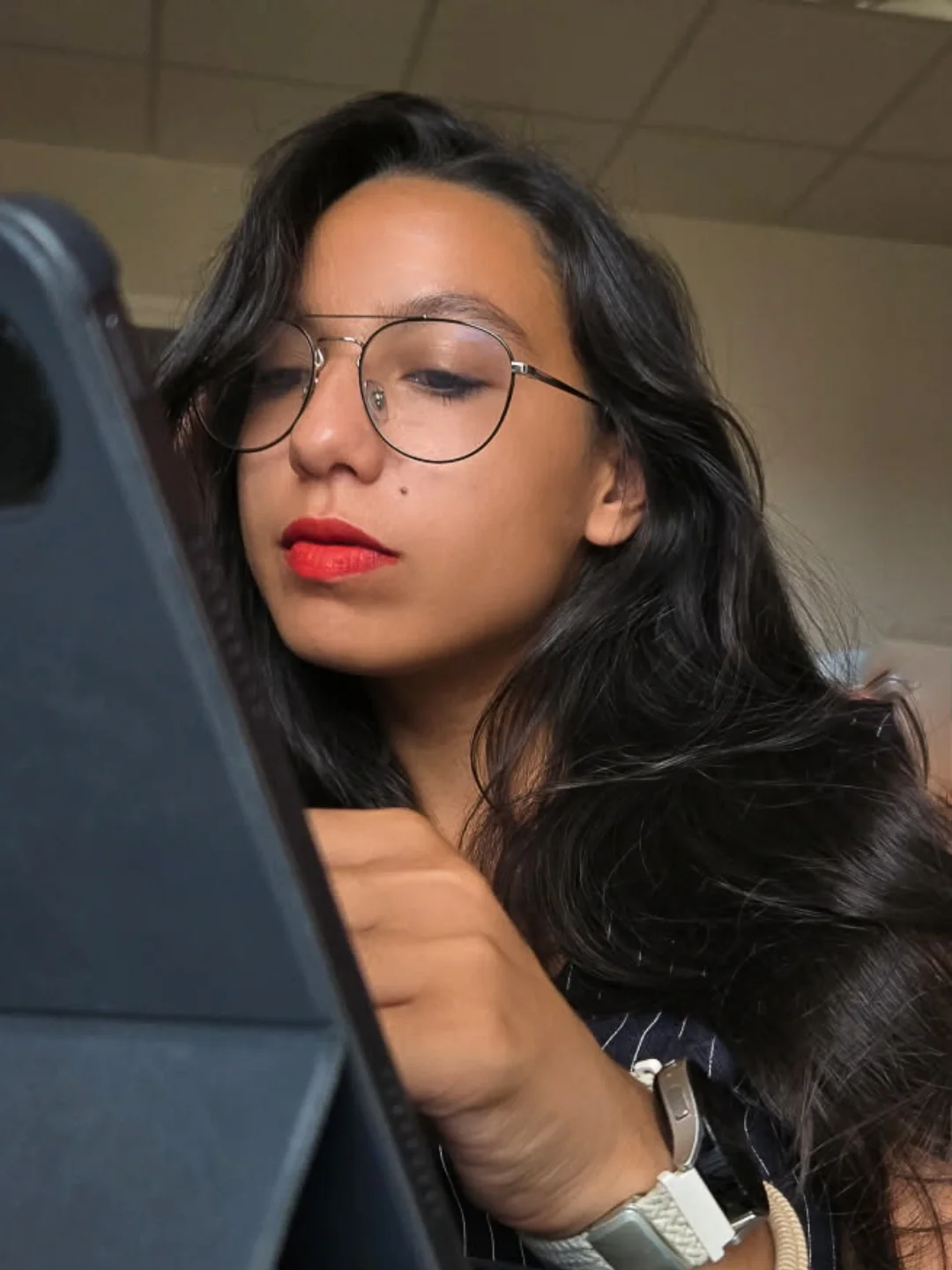 Une jeune femme aux cheveux noirs, portant des lunettes rondes, regarde un téléphone, avec un fond de plafond de bureau.