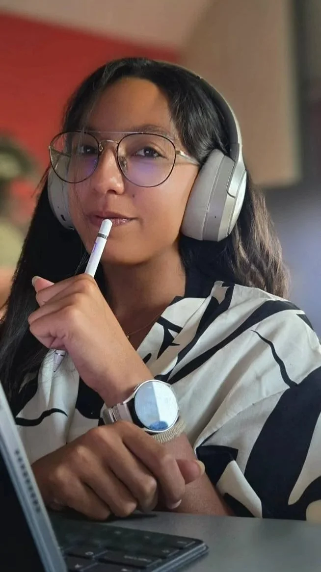 Femme portant un casque audio, portant des lunettes, tenant un stylo près de la bouche, portant une montre et un chemisier à rayures, assise devant un ordinateur.