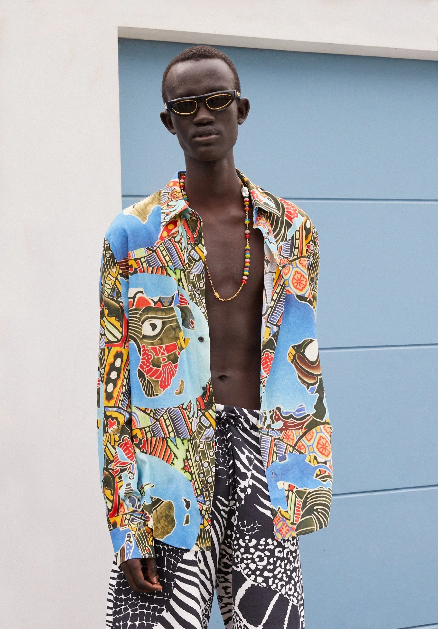 A young man with dark skin and short hair, wearing a colorful patterned open shirt, a beaded necklace, black and white animal print pants, accessorized with sunglasses, standing in front of a white and blue wall.