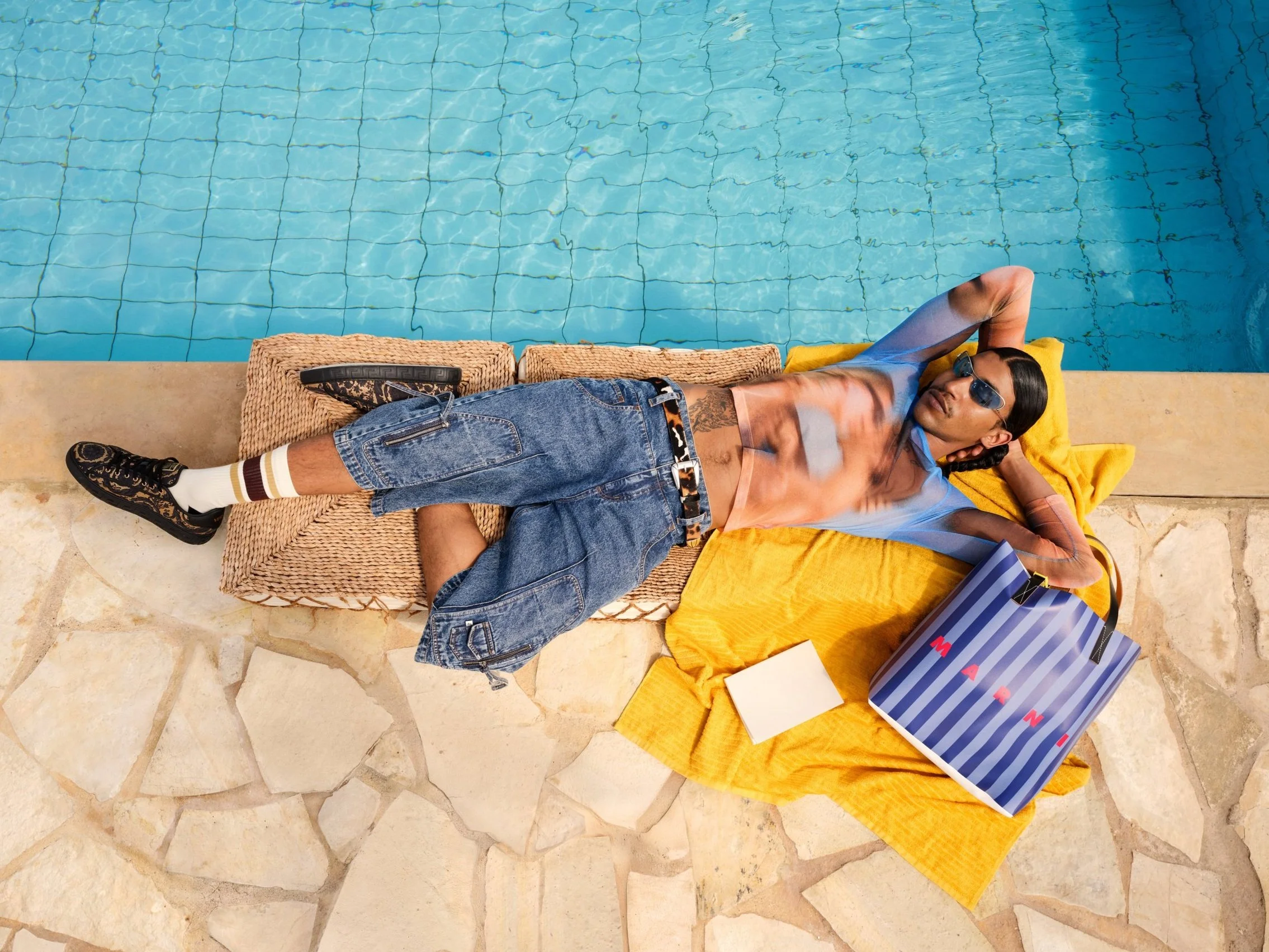 Young man lies by a swimming pool on a towel, wearing sunglasses, a top printed with a blurred photo of two people kissing , and shorts, with a shopping bag that reads Marni nearby.