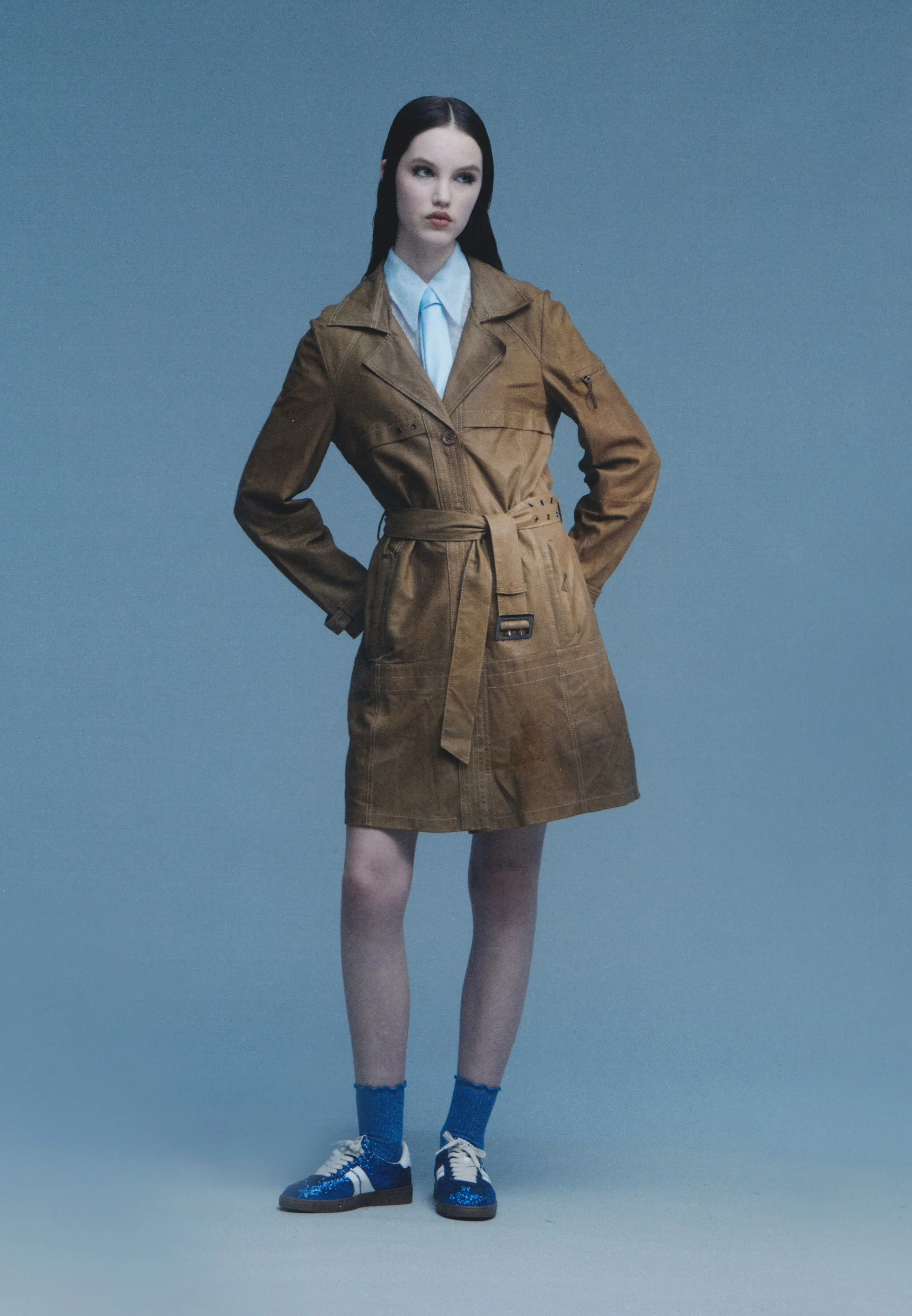 A young woman with long dark hair, wearing a brown trench coat, blue socks, and blue sneakers, stands against a plain gray backdrop with her hands on her hips.