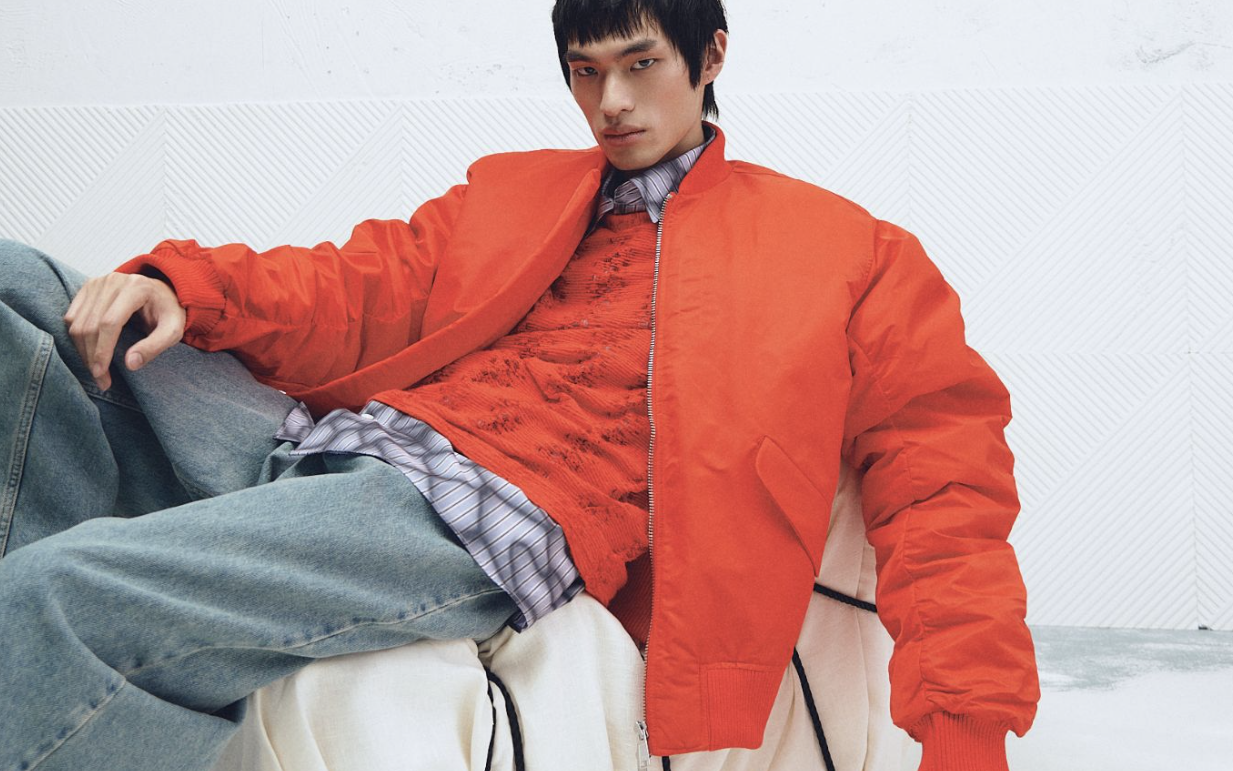 Asian male model in bright orange jacket and knitwear layered over a graphic print button shirt worn with jeans.