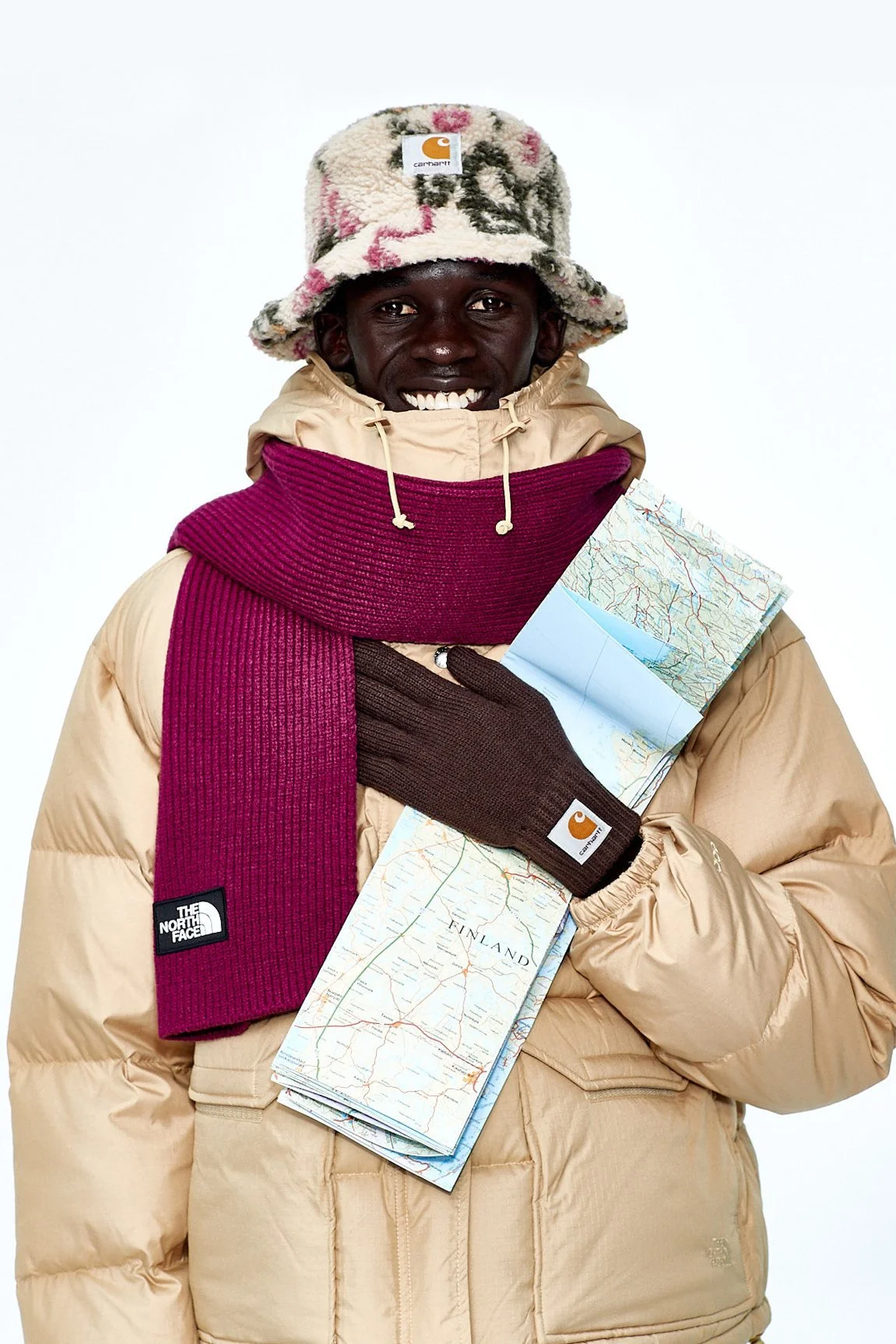 A smiling person dressed warmly in a beige puffy jacket, a colorful fleece hat, a magenta scarf, and brown gloves, holding a folded map.