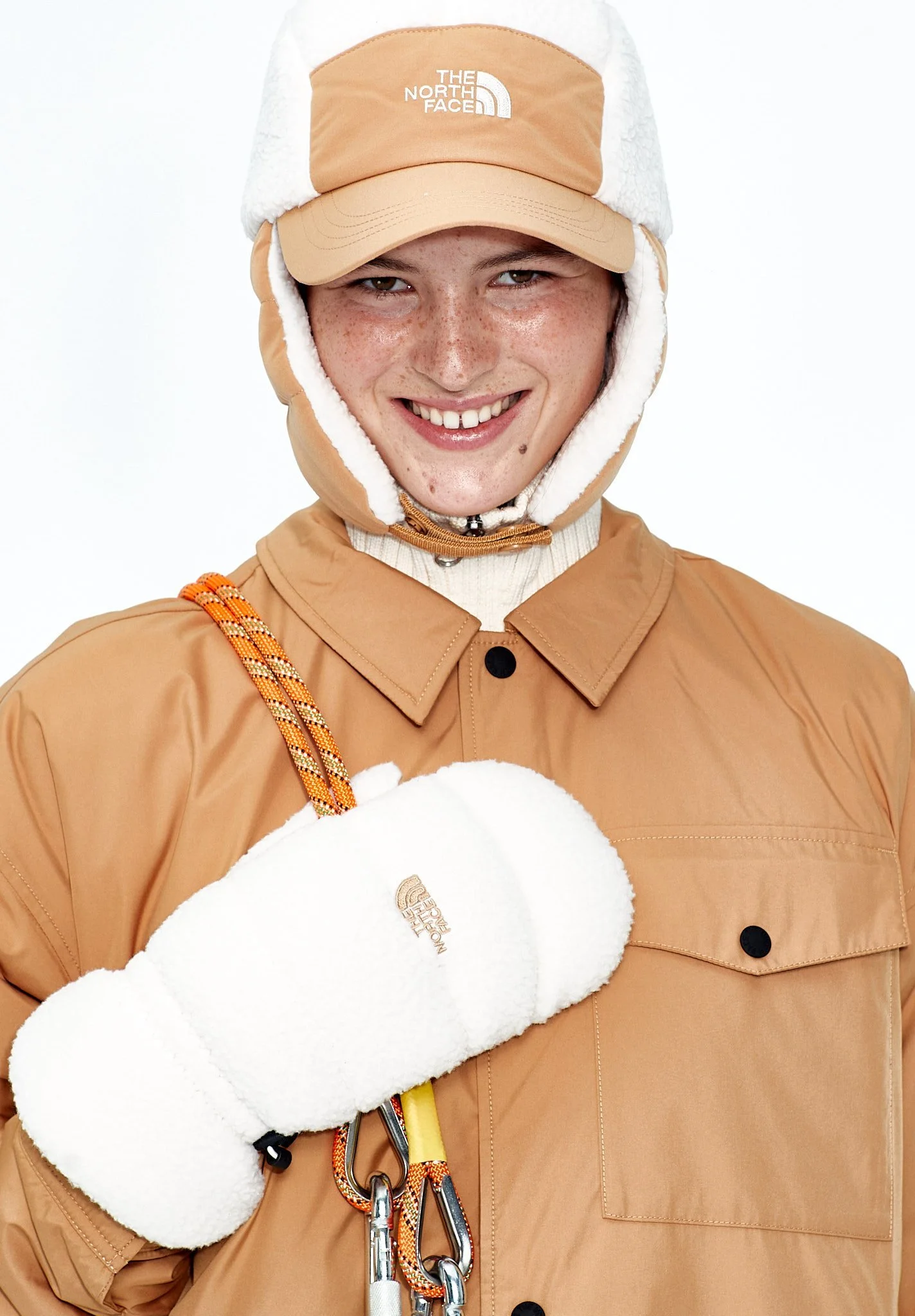 A smiling young woman dressed in beige outdoor gear, including a fleece-lined hat, jacket, and gloves, with climbing clips hanging from her shoulder.