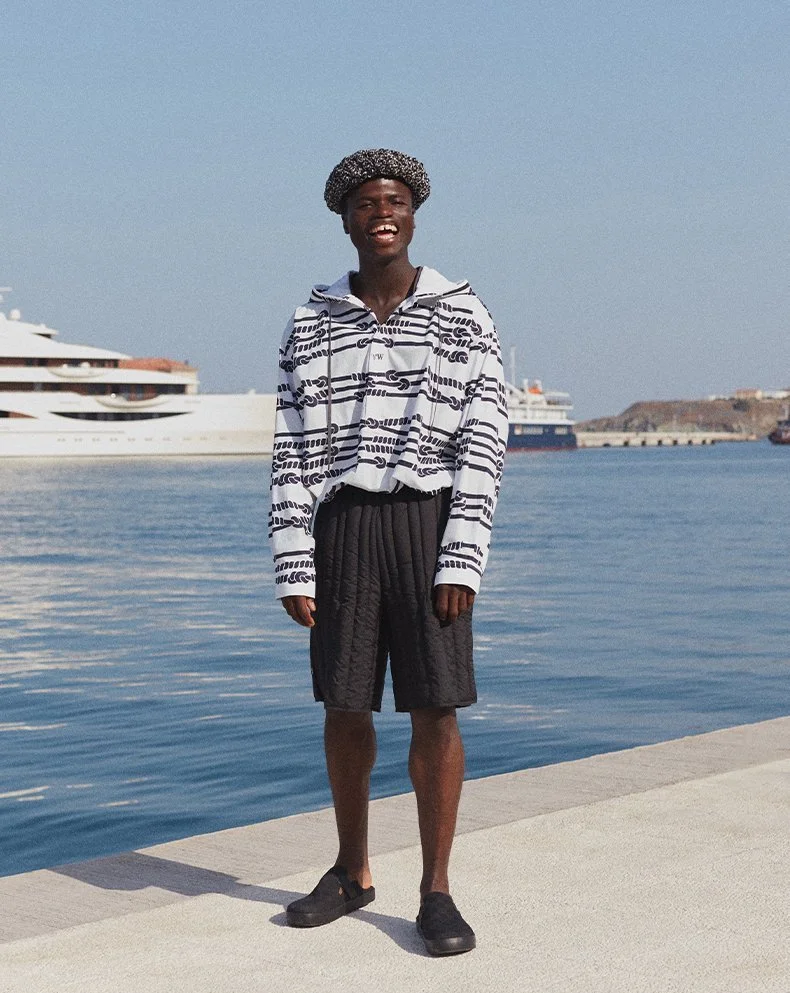 A smiling young man standing on a dock near the water with large yachts in the background, wearing a patterned hoodie, black shorts, and sneakers.