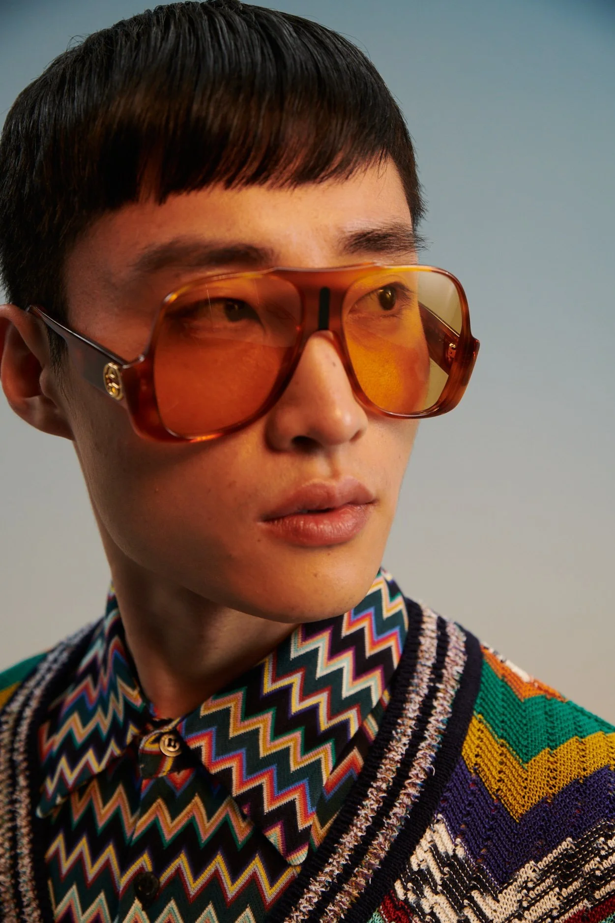 Close-up of a young man with short dark hair wearing large amber sunglasses and a colorful zigzag patterned shirt with a multicolored knitted sweater.