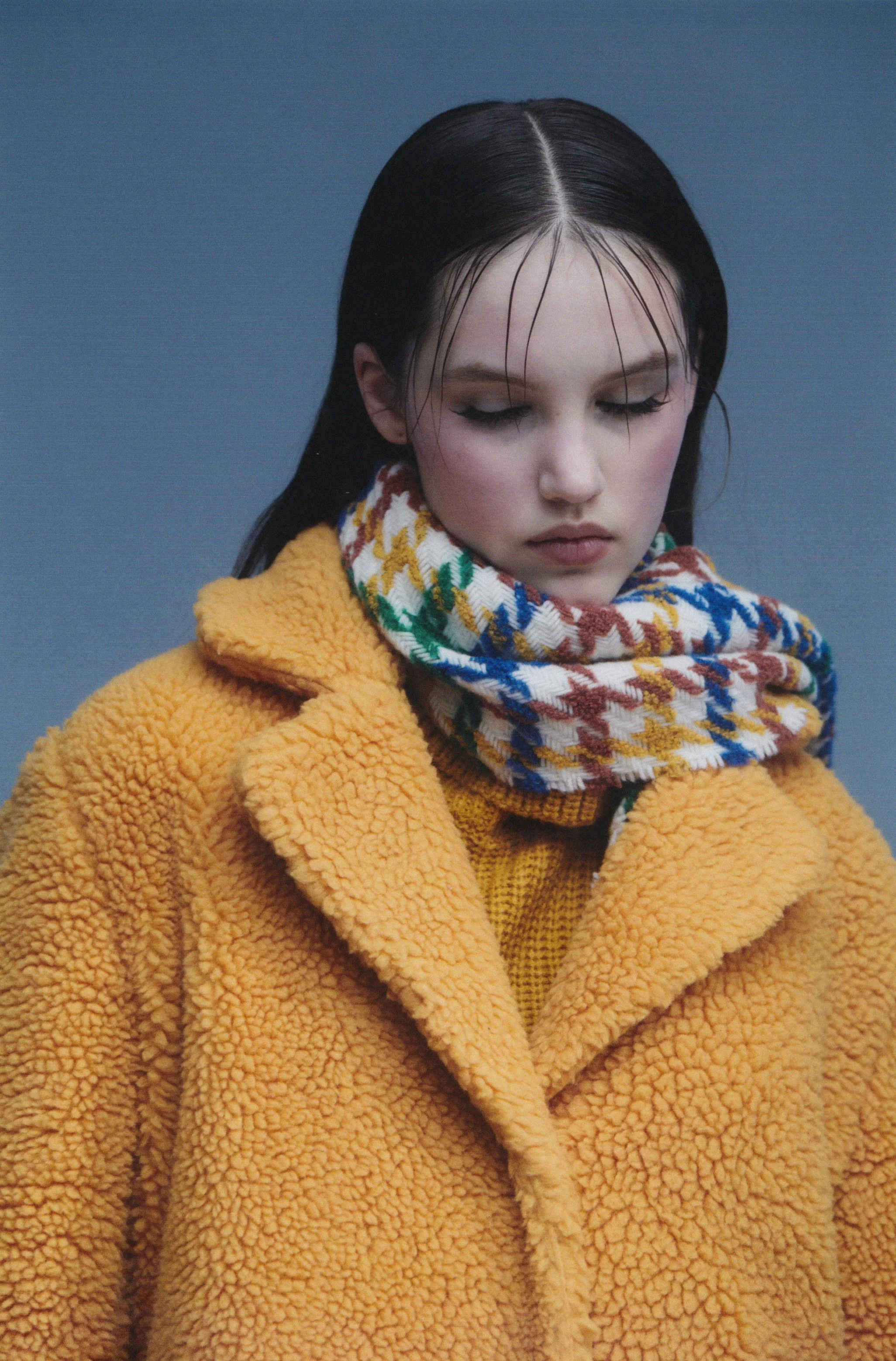 A young woman with wet, slicked-back black hair wearing a yellow fuzzy coat and a multicolored plaid scarf against a blue background.
