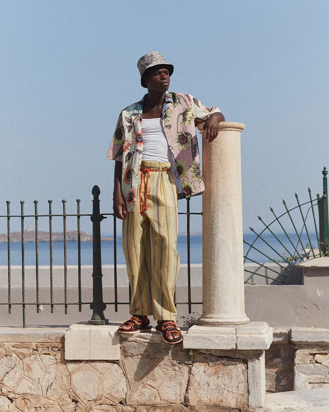 A young man standing outdoors against a clear blue sky, wearing a floral shirt, striped pants, and a bucket hat, with a column and ocean in the background.