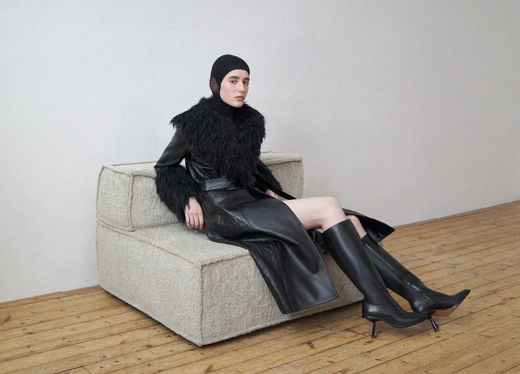 A woman dressed in a black leather outfit with knee-high black boots, sitting on a beige textured sofa against a plain wall with wooden flooring.