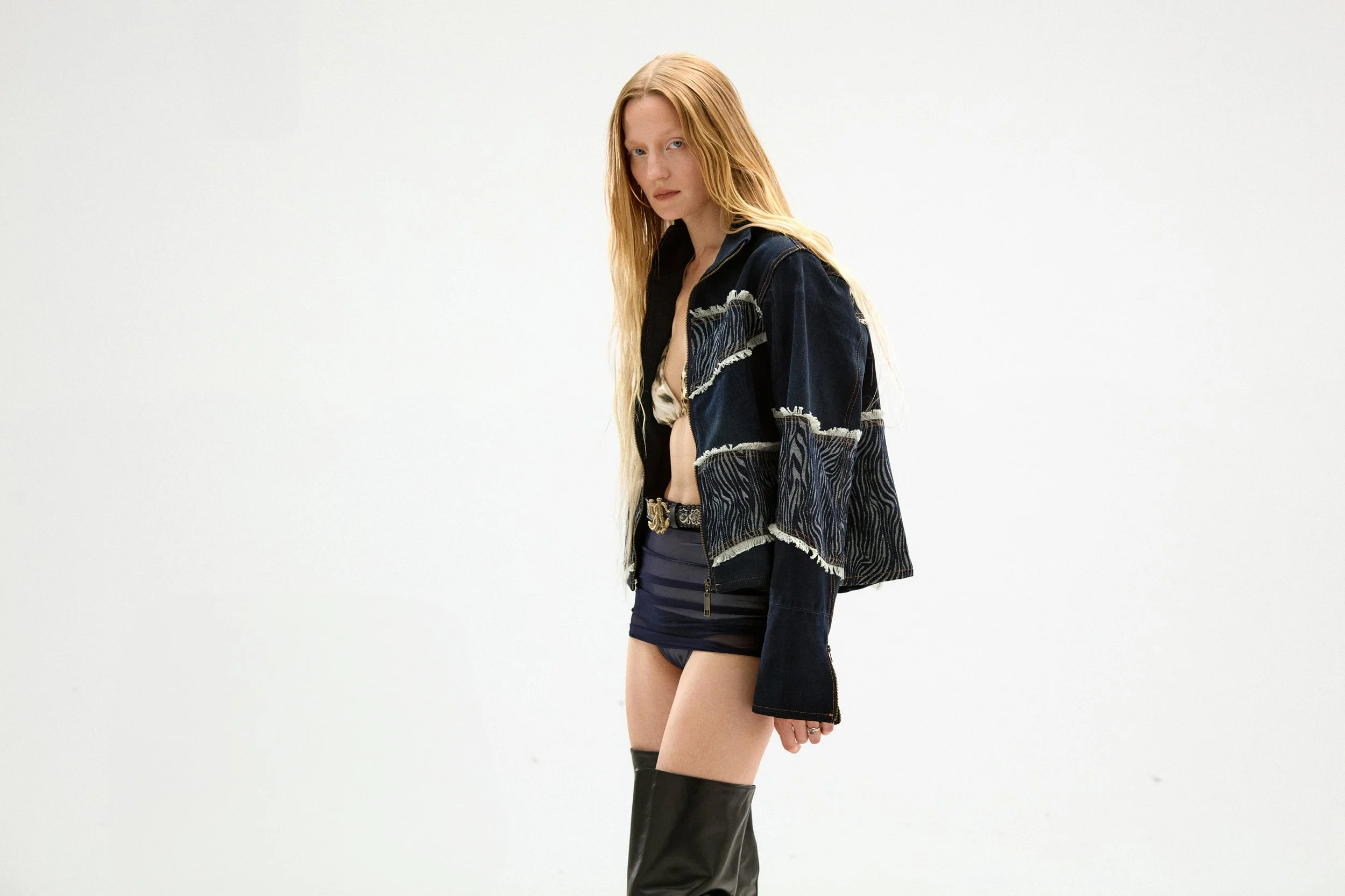 Young woman with long dark blonde hair wearing a patterned black and blue jacket, a revealing top, a belt, and thigh-high black boots, standing against a plain white background.