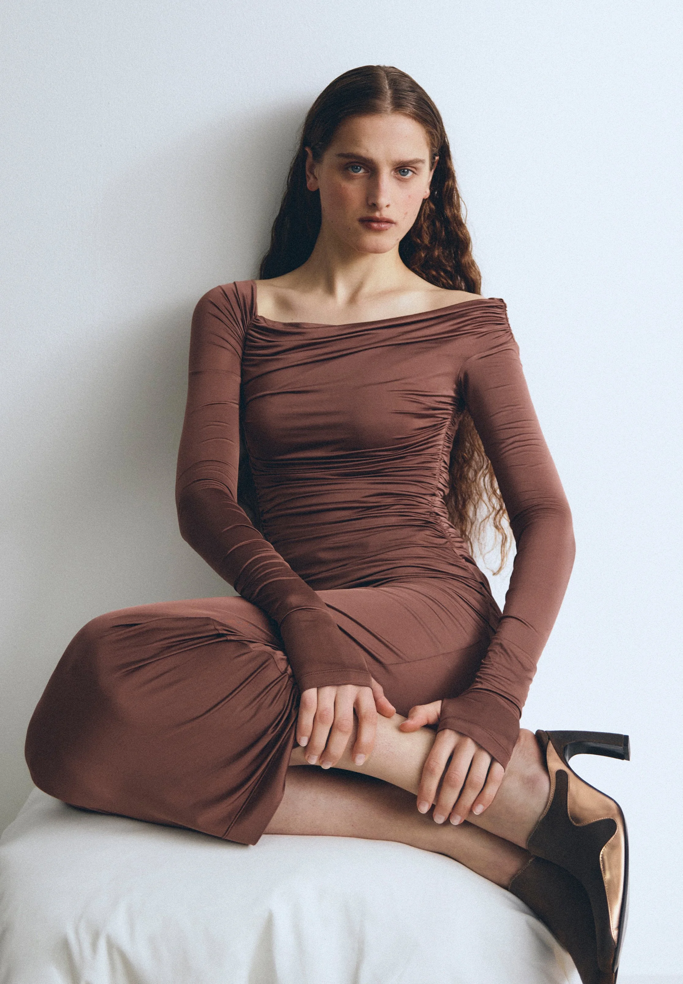 A woman with long wavy hair sitting on a white surface, wearing a brown long-sleeved, ruched dress and dark heels, against a plain white wall.