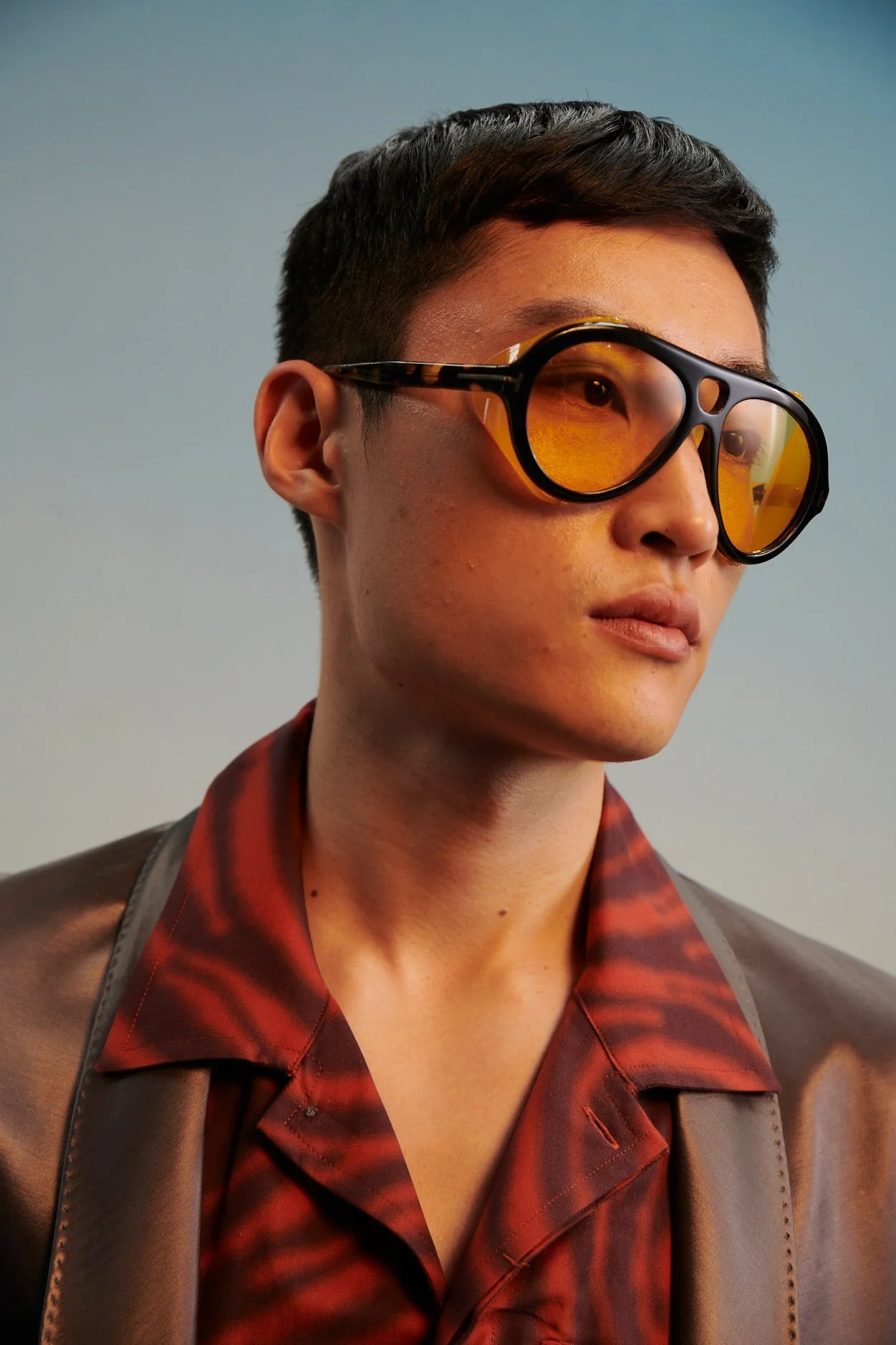 A young man wearing yellow-tinted sunglasses, a metallic brown jacket, and a red and black patterned shirt against a plain background.