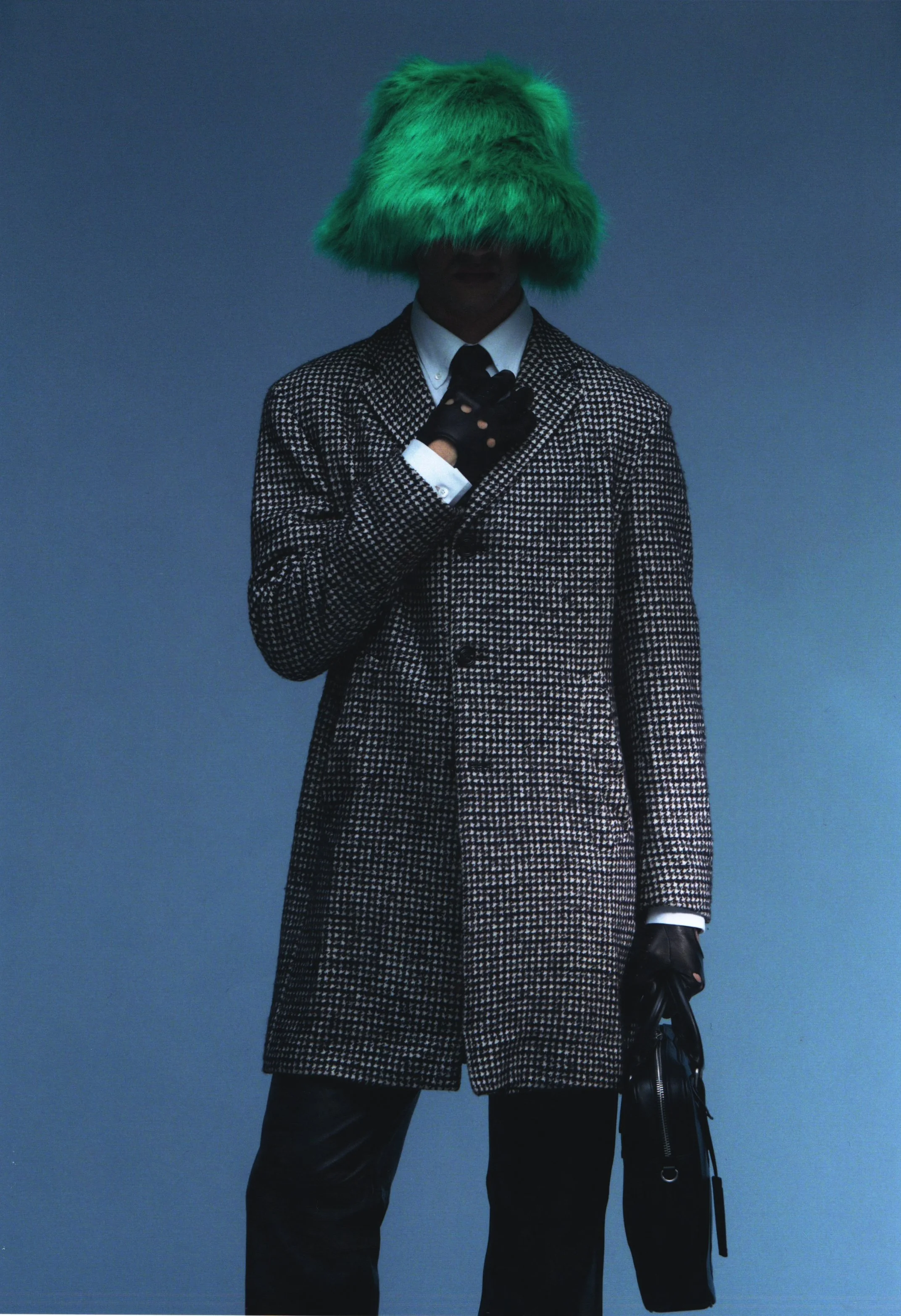 Person wearing a green fuzzy hat, a black and white checkered coat, black gloves, holding a black bag, against a plain blue background.