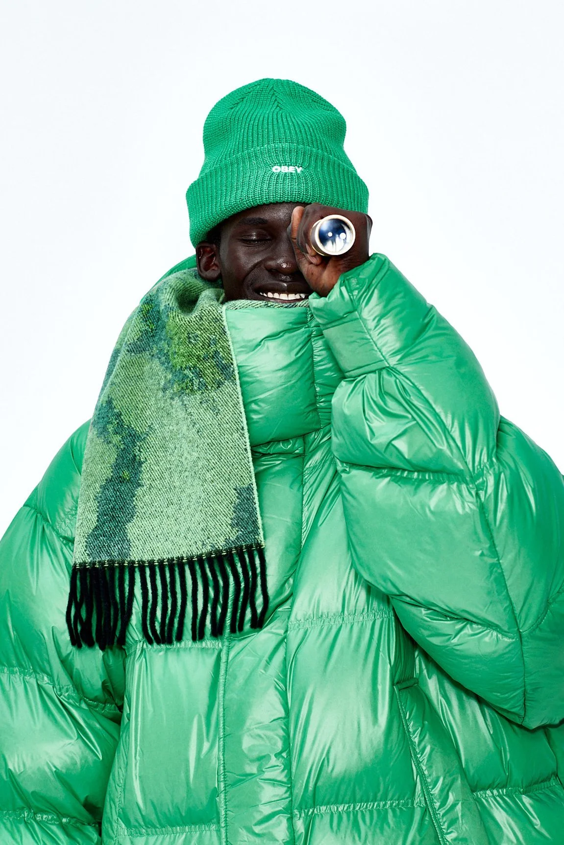 Person wearing a green beanie, green puffer jacket, and a green scarf with black fringe, holding a telescope up to their eye as if viewing something in the distance.