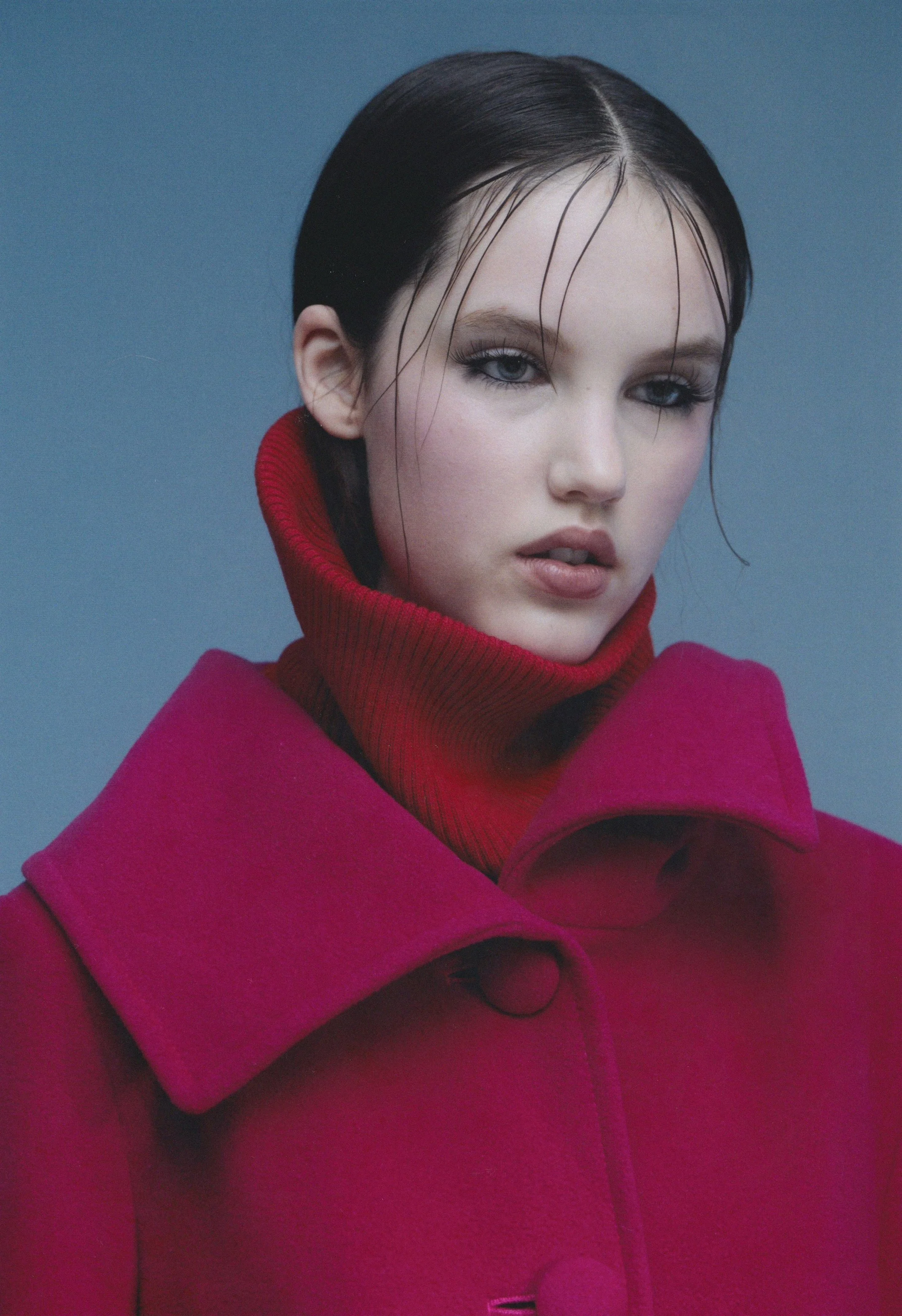 A young woman with wet dark hair slicked back, wearing a red turtleneck and a magenta coat with a large collar, standing against a blue background.
