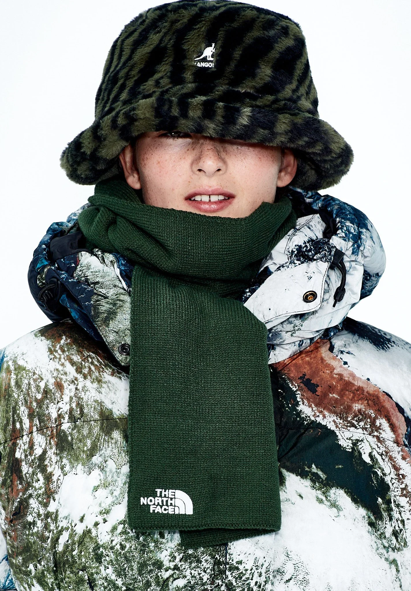 Person wearing a dark and green furry hat, a green scarf, a patterned snow jacket, and a green The North Face scarf.