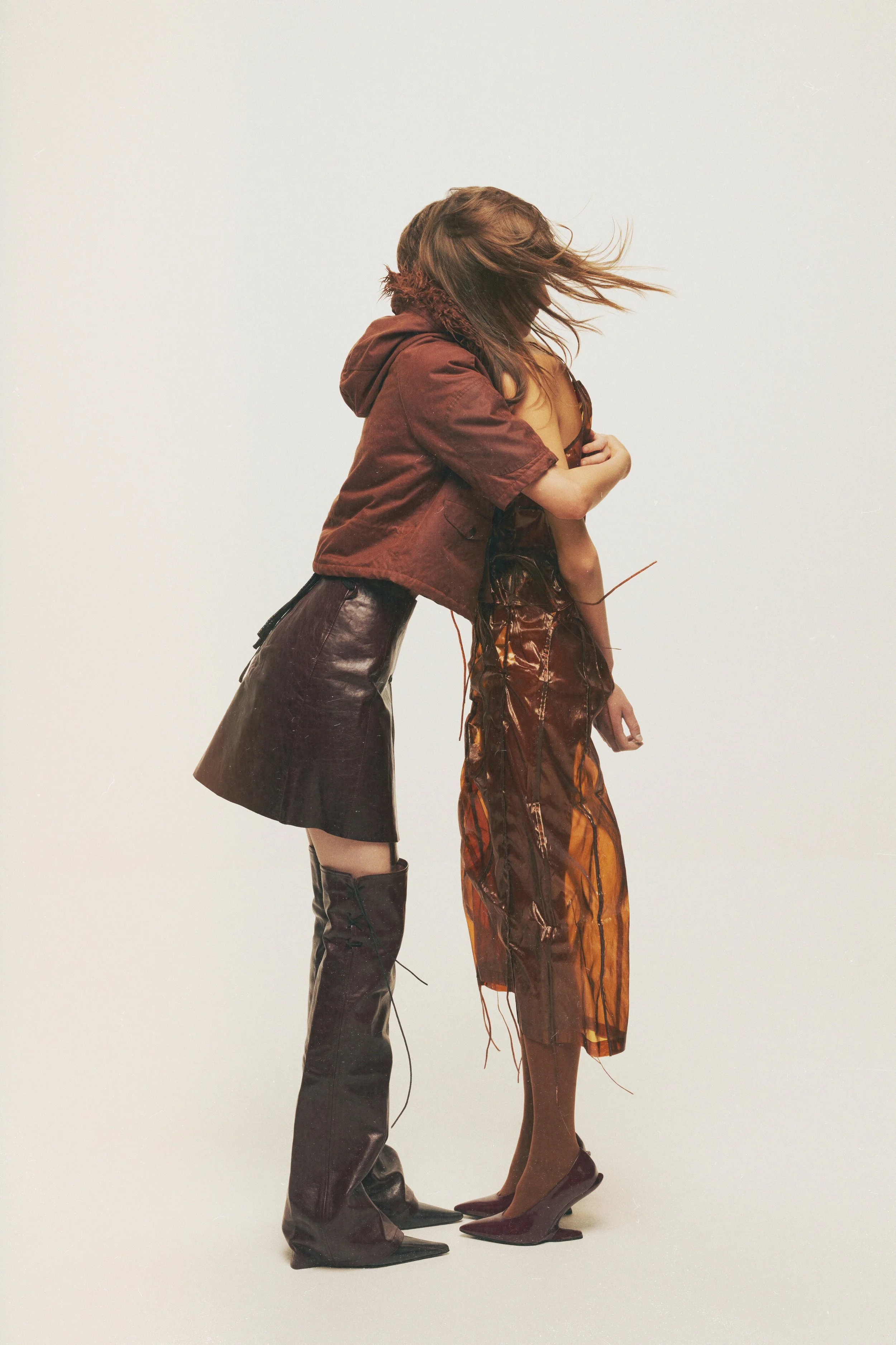 Two women standing closely, one leaning over the other, with the woman on the right appearing to be in a distressed state. Both are dressed in brown-toned clothing, with the woman on the left wearing a jacket and long pants, and the woman on the righ