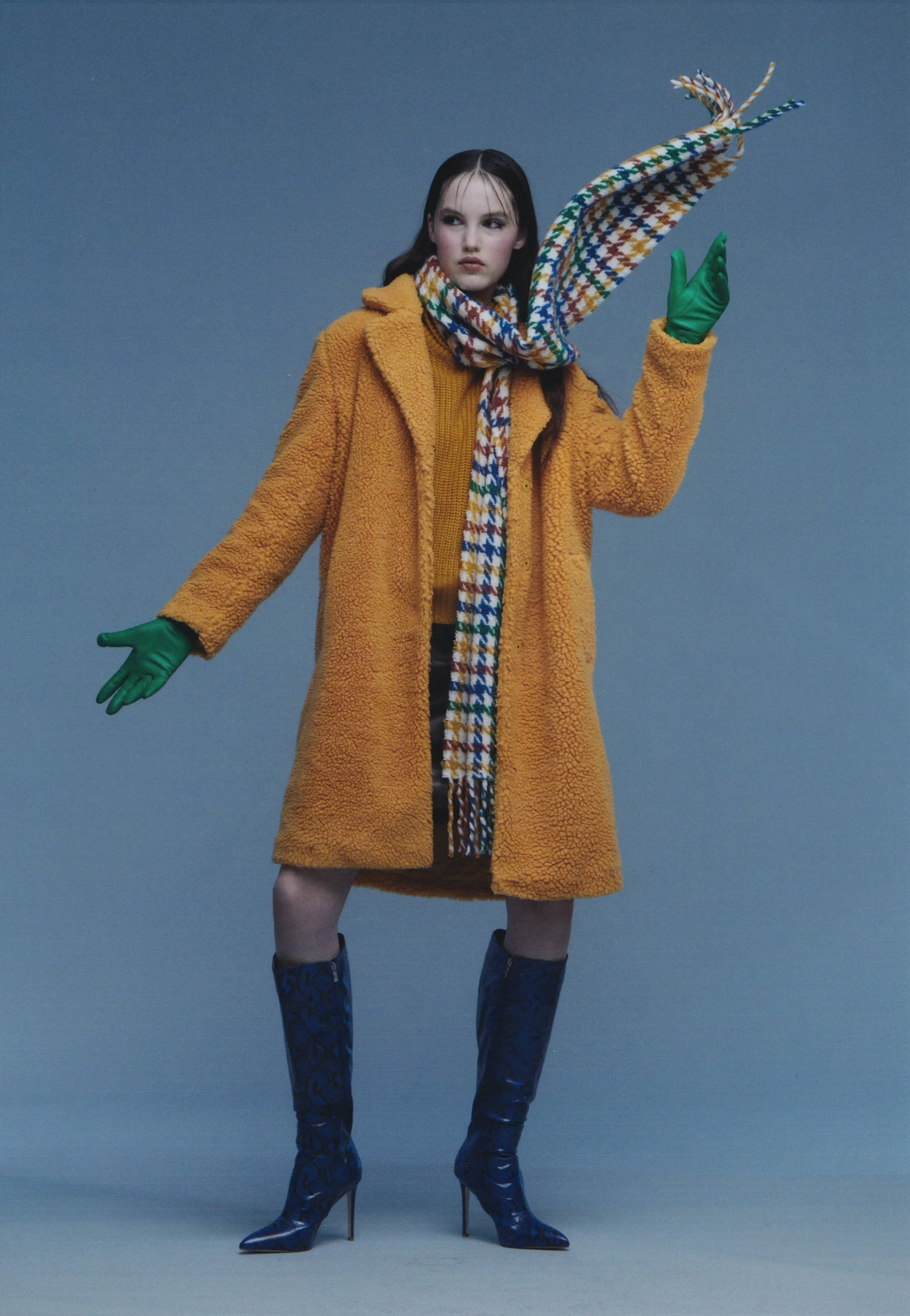 A woman standing against a powdery blue background, wearing a yellow coat, a colorful scarf, green gloves, blue high-heeled boots. She stands in a dynamic pose as her scarf is in motion.