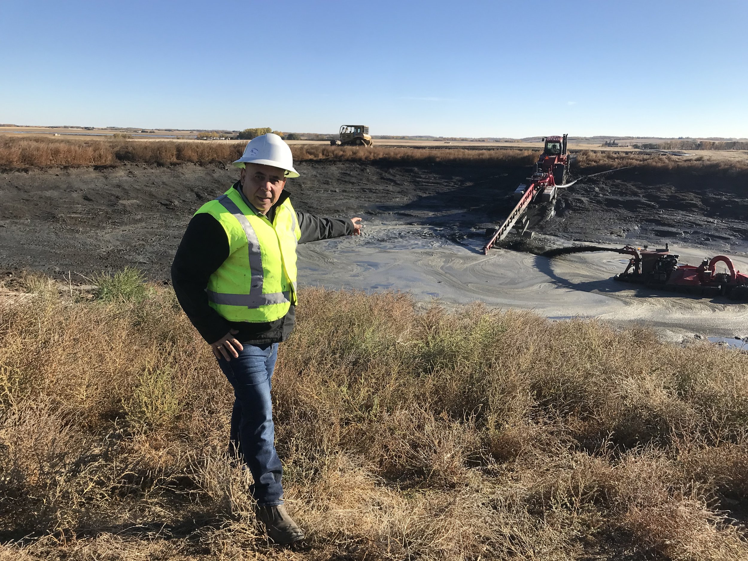 A man wearing a white safety helmet and neon yellow safety vest standing on dry grass and pointing towards a burned area in a field with a large soil stabilization or removal machine in the background.