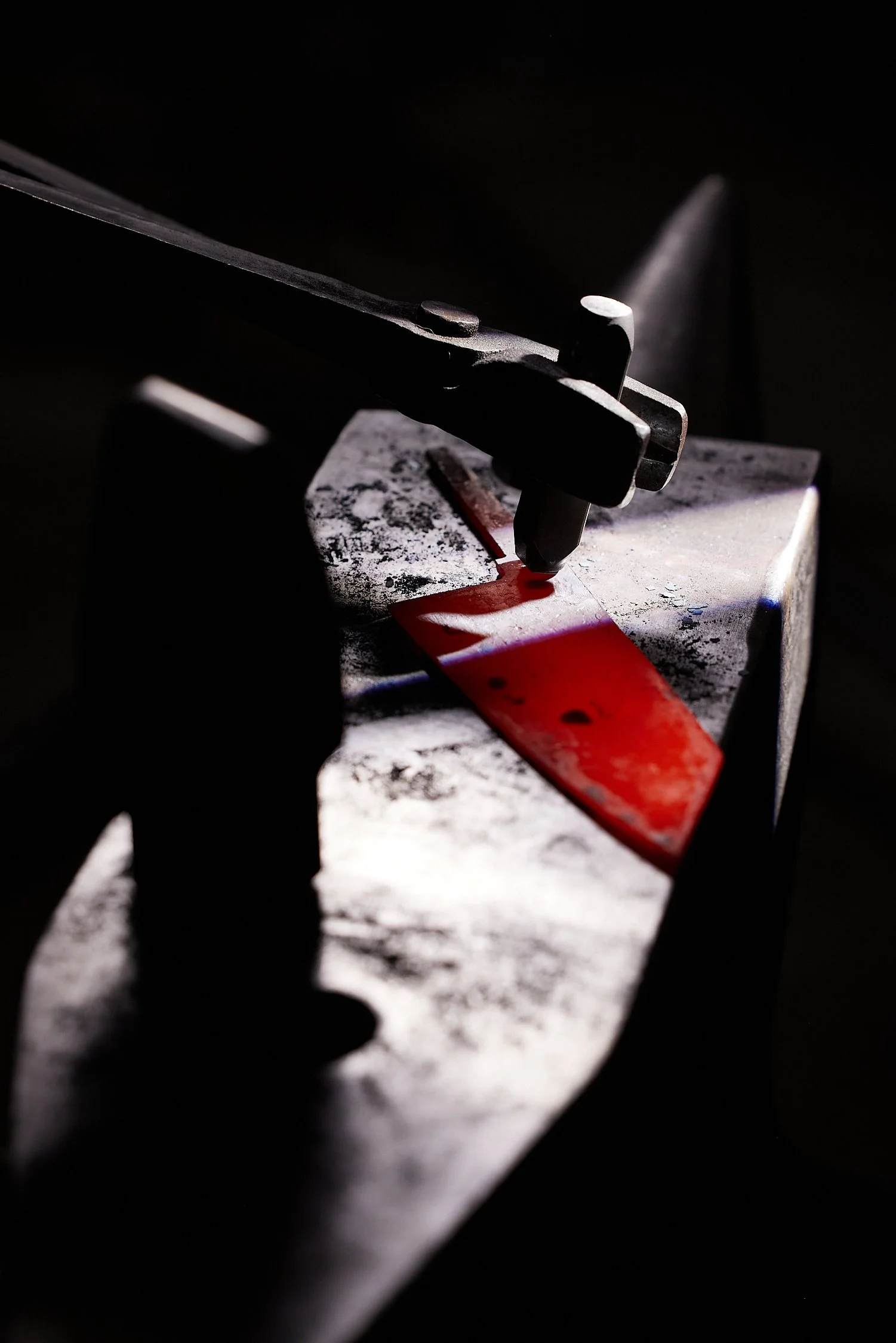 A blade is being shaped on a grinding wheel with a red-and-black handle and black clamp in a dark workshop.