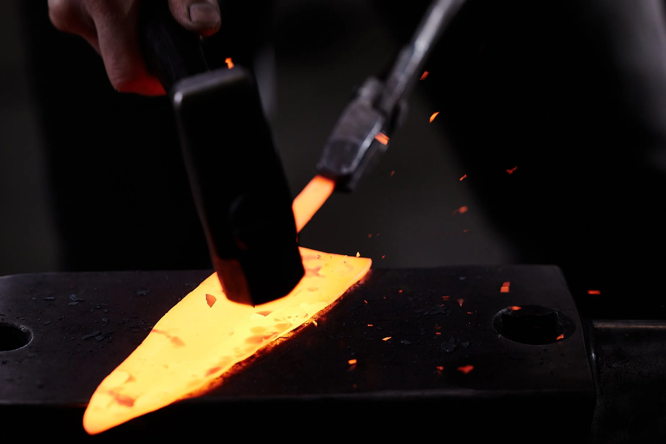 A blacksmith forging metal on an anvil with sparks flying, using a hammer and tongs in a dark workshop.