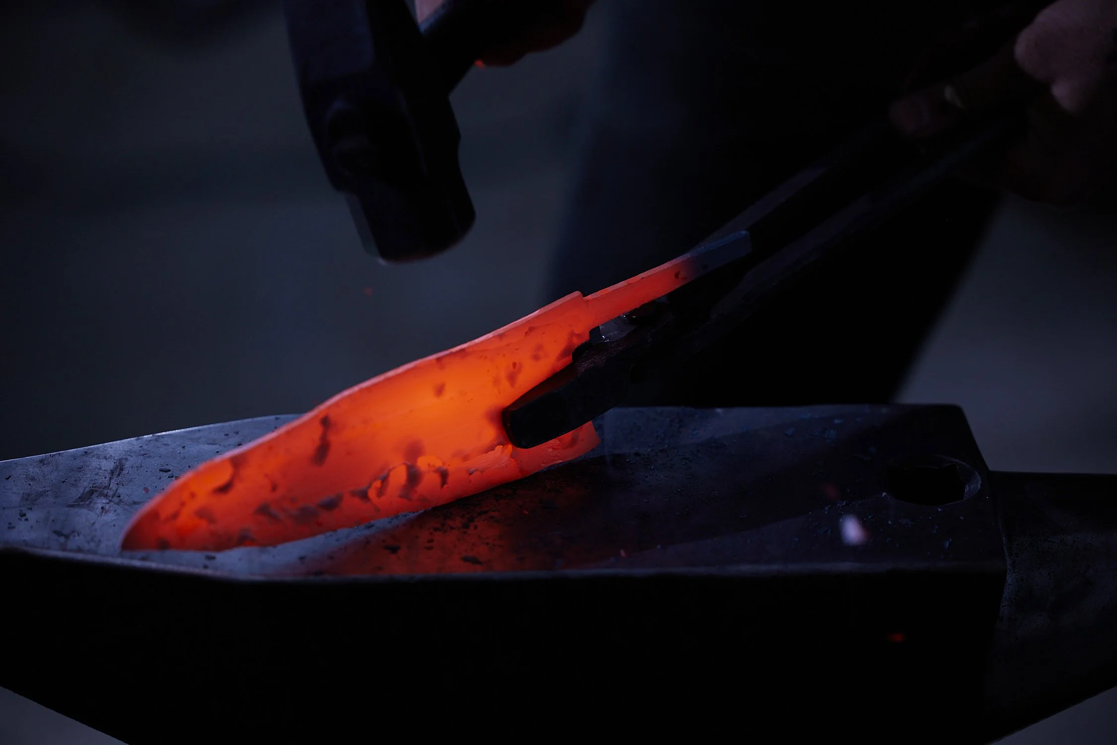 A blacksmith shaping glowing hot metal on an anvil with a hammer in a dark workshop.