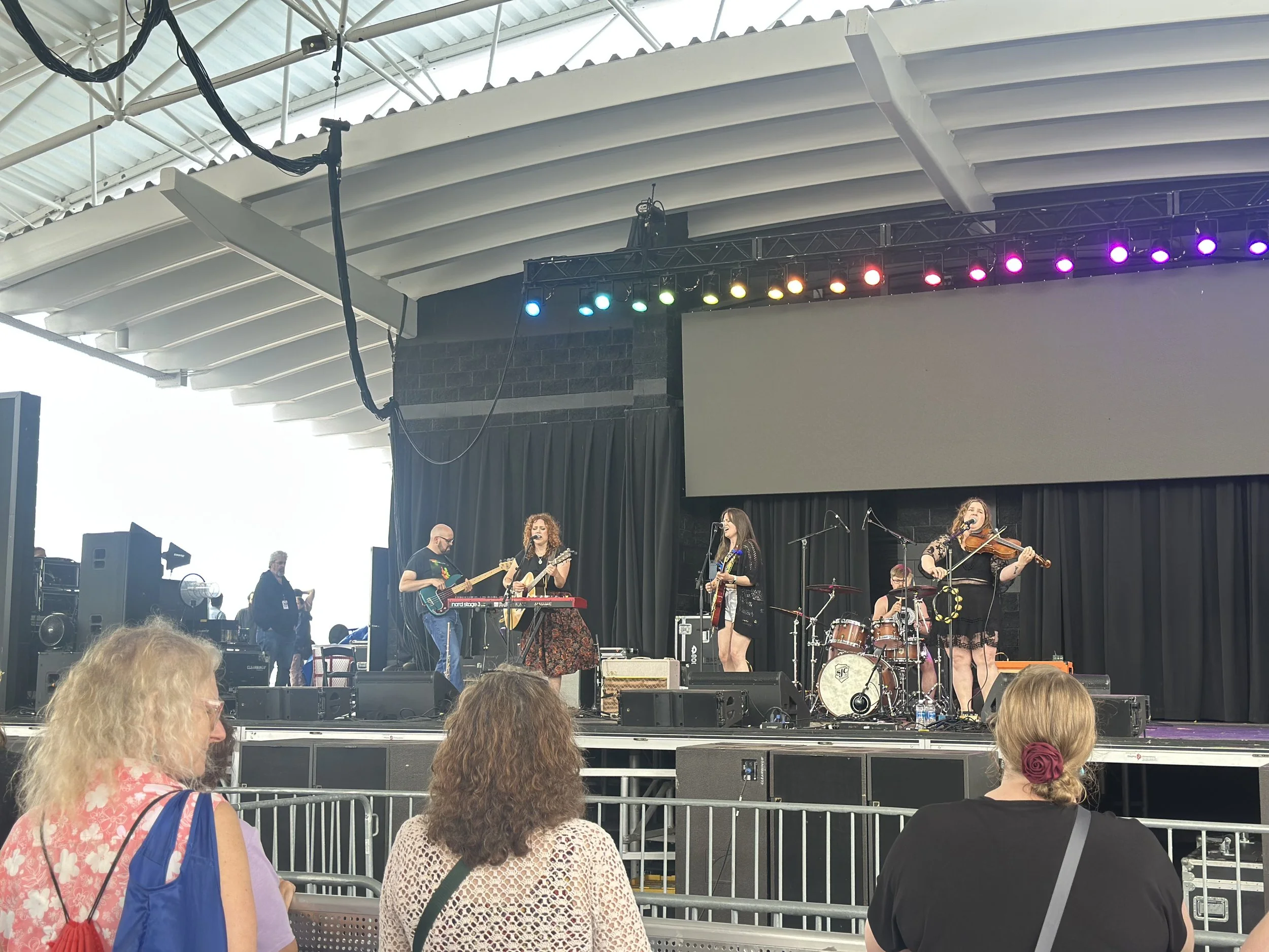 A band performs live on stage at a concert venue with an audience watching. The stage has musical instruments and colorful lighting.