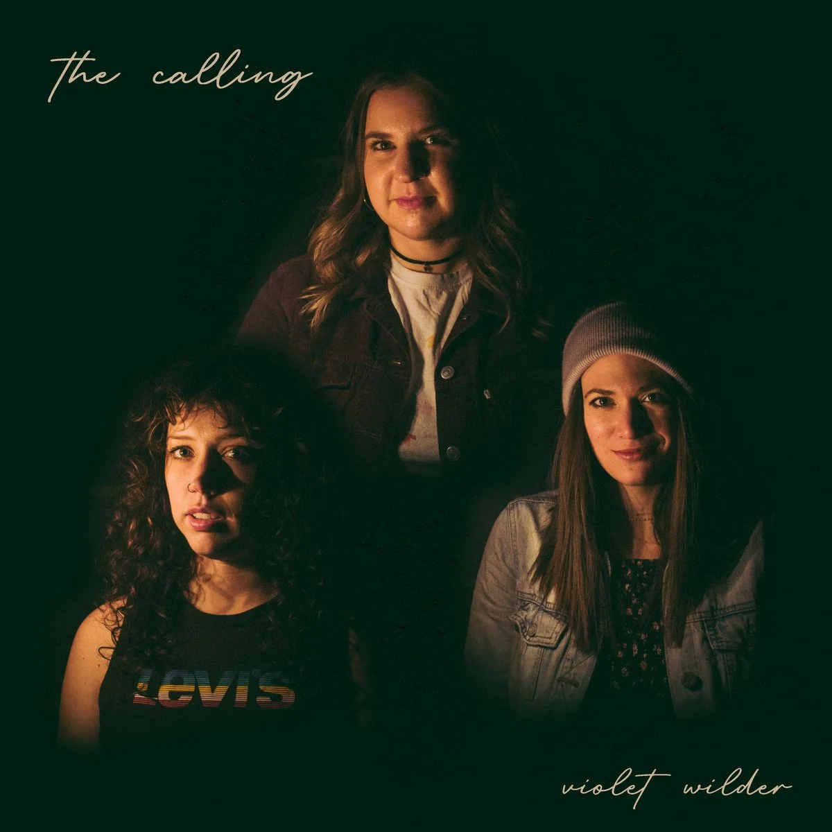 A promotional image featuring three women, with the text 'the calling' in the top left and 'violet wilder' in the bottom right. The women have different hairstyles and outfits, and are set against a dark background.