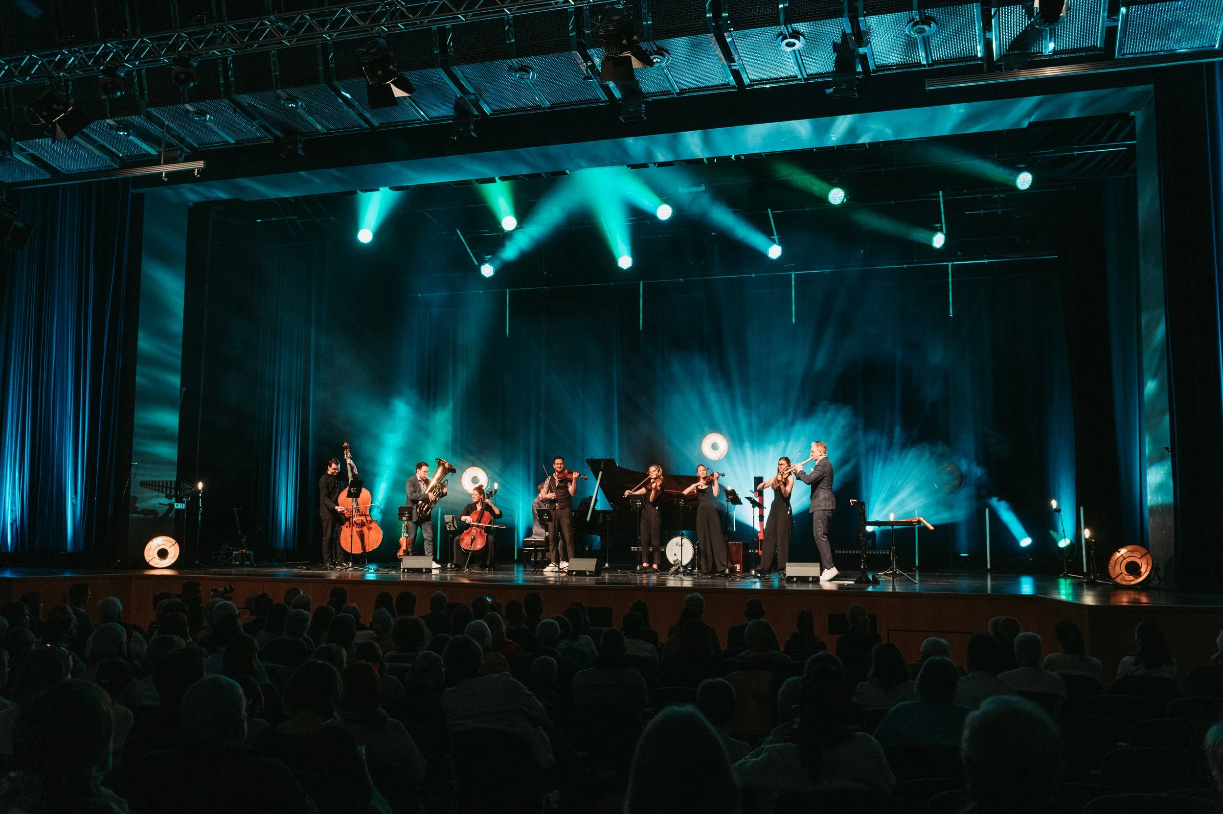 Orchester auf einer Bühne mit blauer Beleuchtung, bestehend aus Musikern, die verschiedene Instrumente spielen, vor einem dunklen Hintergrund, während ein Publikum im Saal zuschaut.