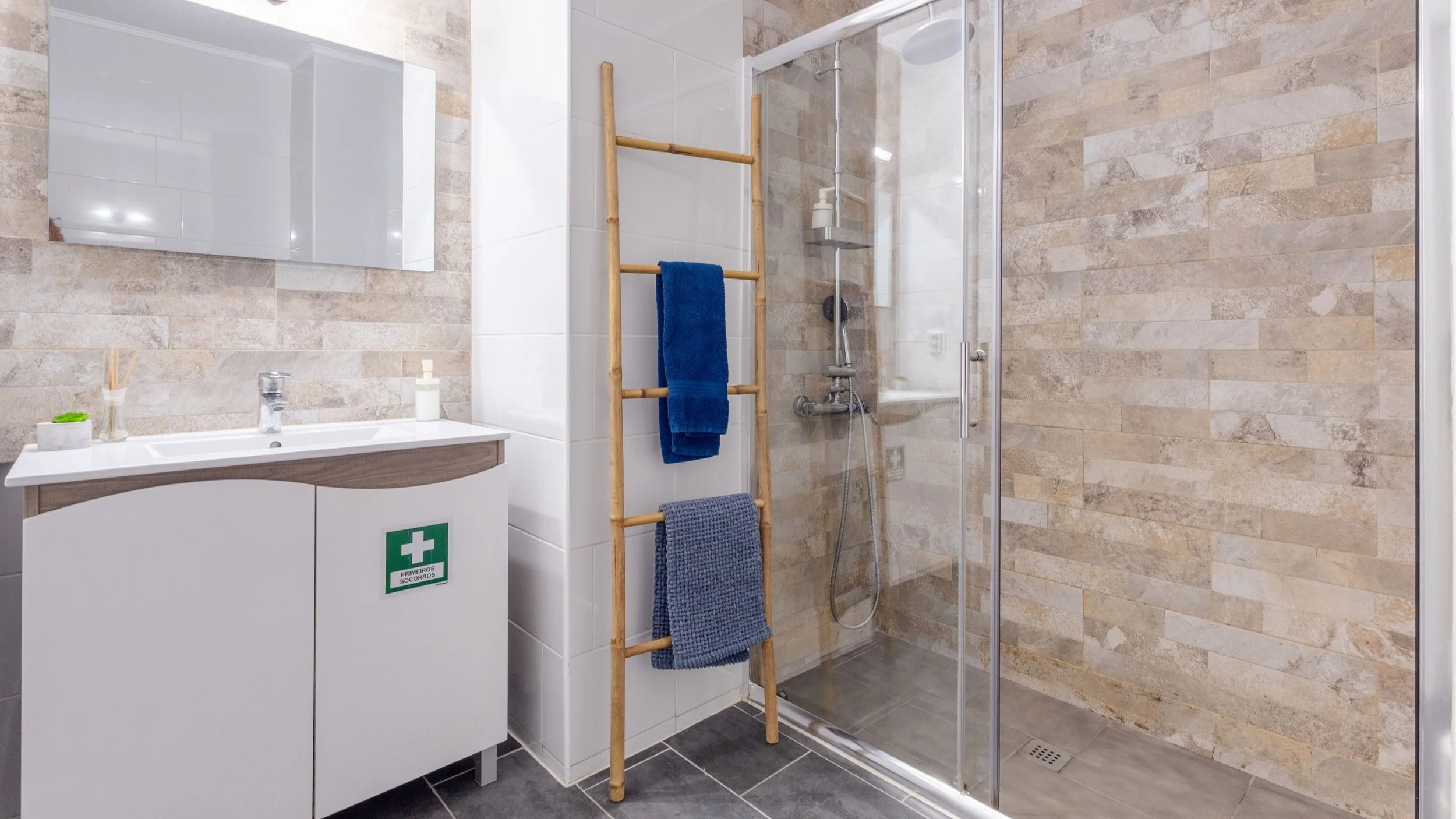 Banheiro com pia, espelho, cabideiro de madeira com toalhas, box de vidro com ducha, parede de azulejos bege, piso preto, e produtos de higiene.