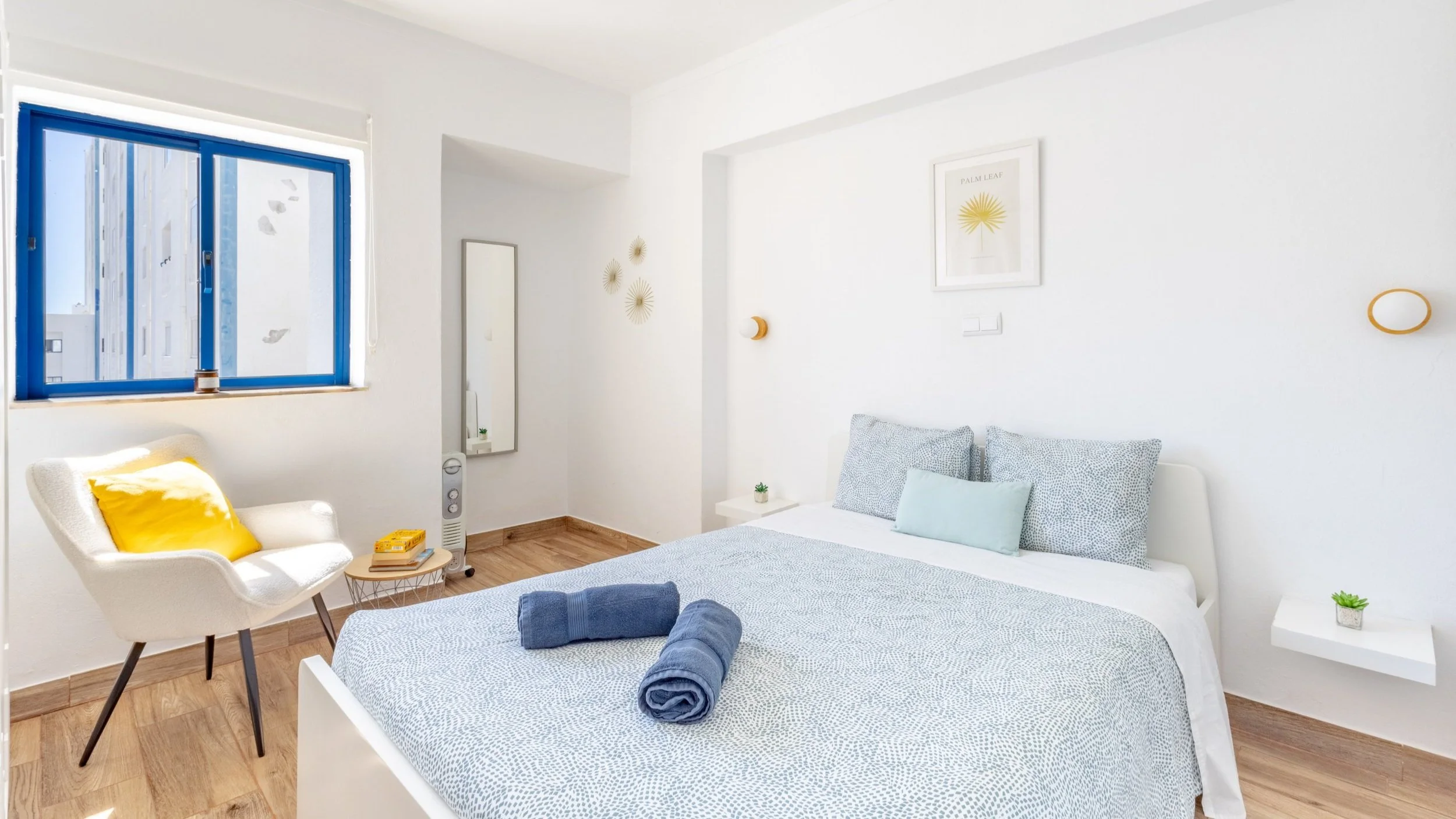 Quarto com cama de casal, roupas de banho azul, parede branca, janelas com moldura azul, cadeira branca com almofada amarela, decoração minimalista com quadros e plantas pequenas.