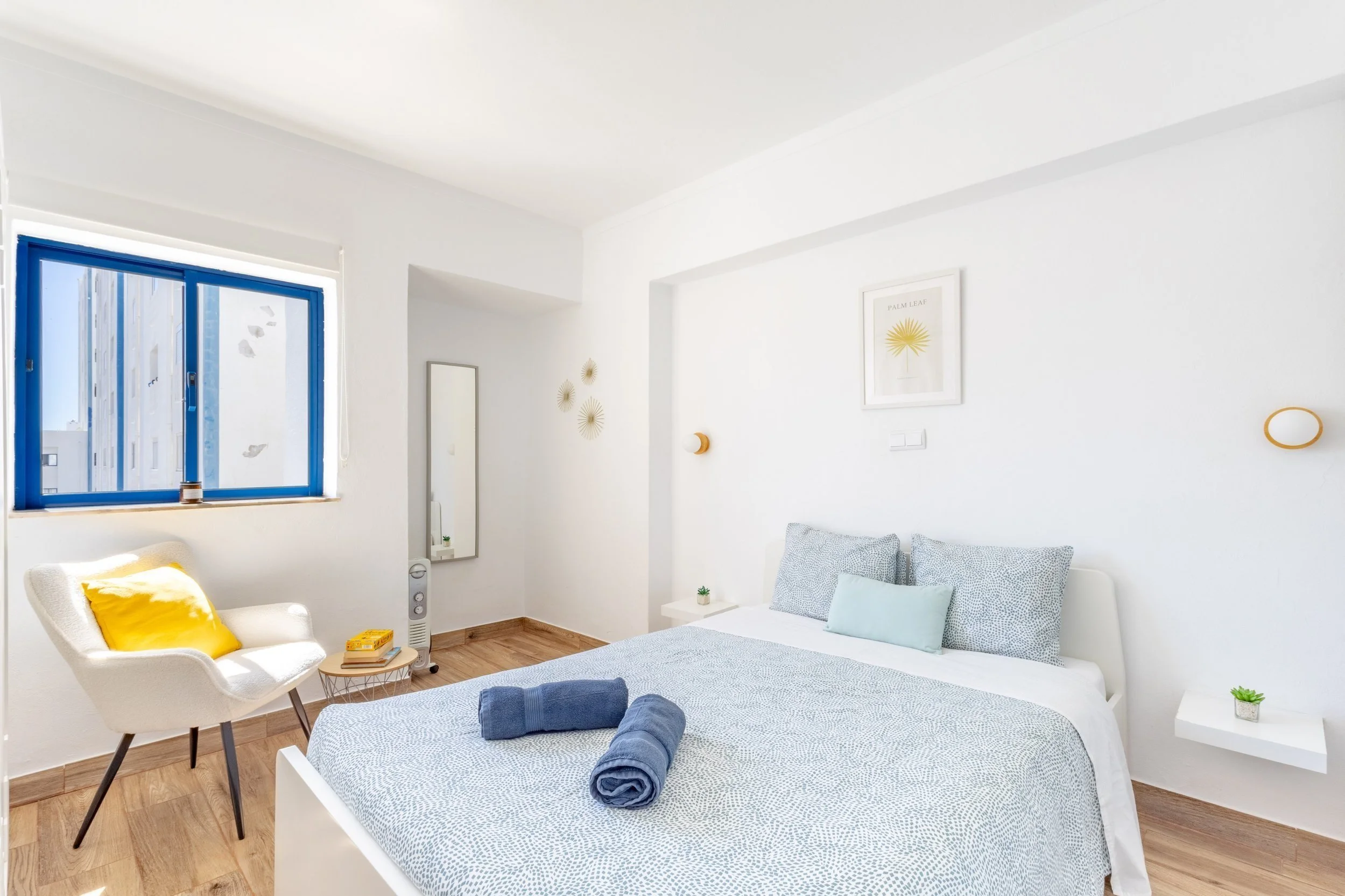 Quarto de casal com paredes brancas, cama com roupa de cama em tons de cinza e azul, duas janelas com moldura azul, cadeira branca com almofada amarela, e detalhes decorativos na parede.