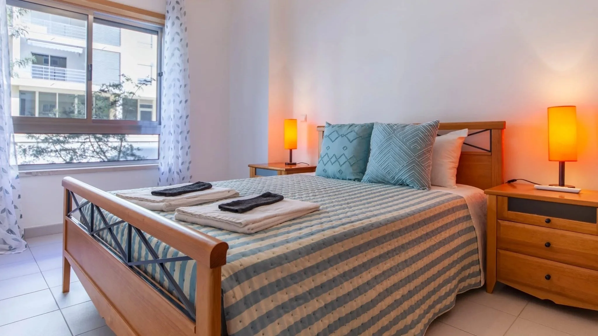 Quarto com cama de madeira, roupas dobradas na cama, duas almofadas azuis e duas branquinhas, luminárias acesas ao lado da cama, janela com cortinas brancas com detalhes azuis, teto de vidro mostrando edifícios ao redor.