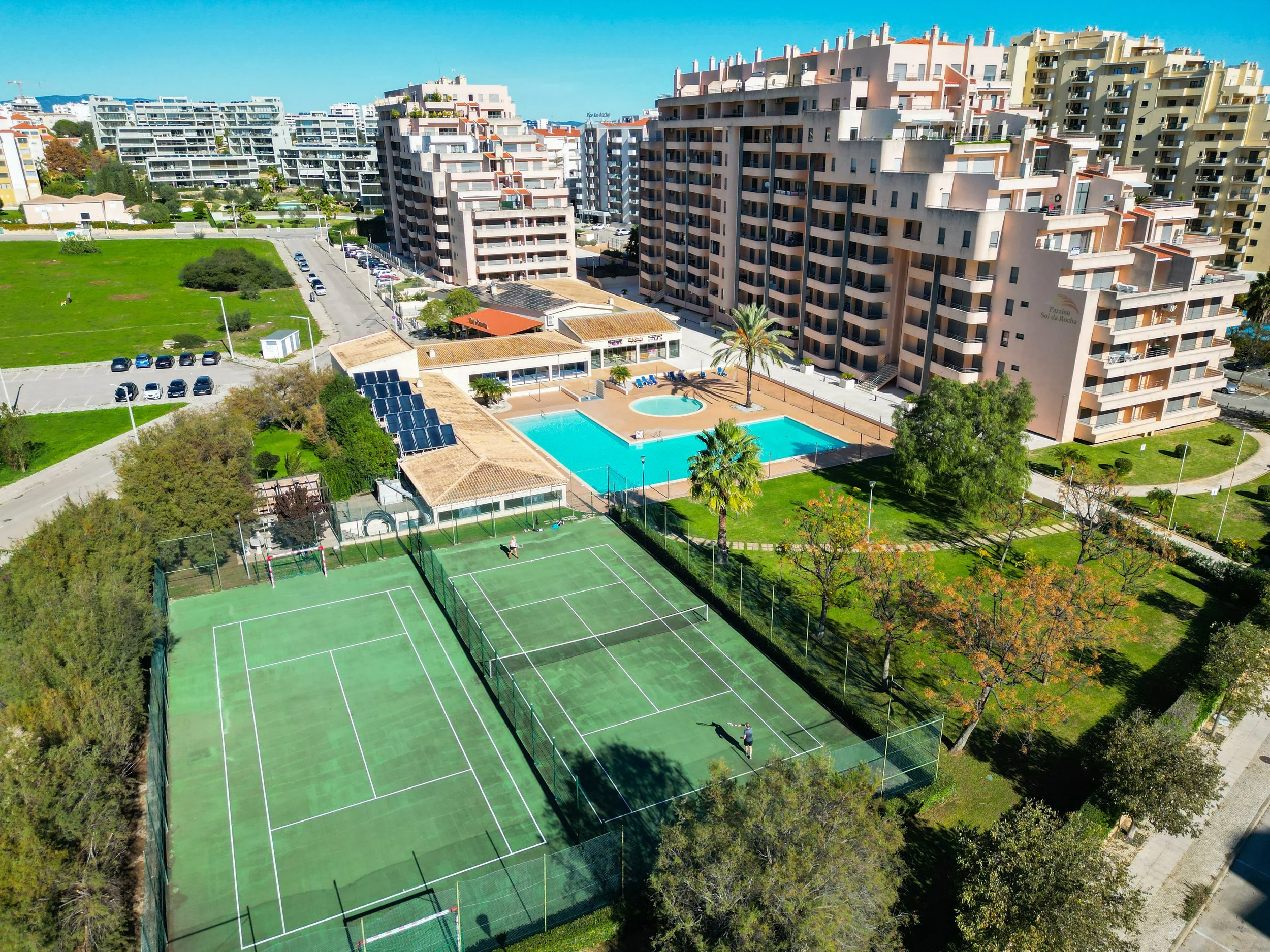 Condomínio com quadras de tênis, piscina, área de lazer com árvores e prédios residenciais ao fundo.