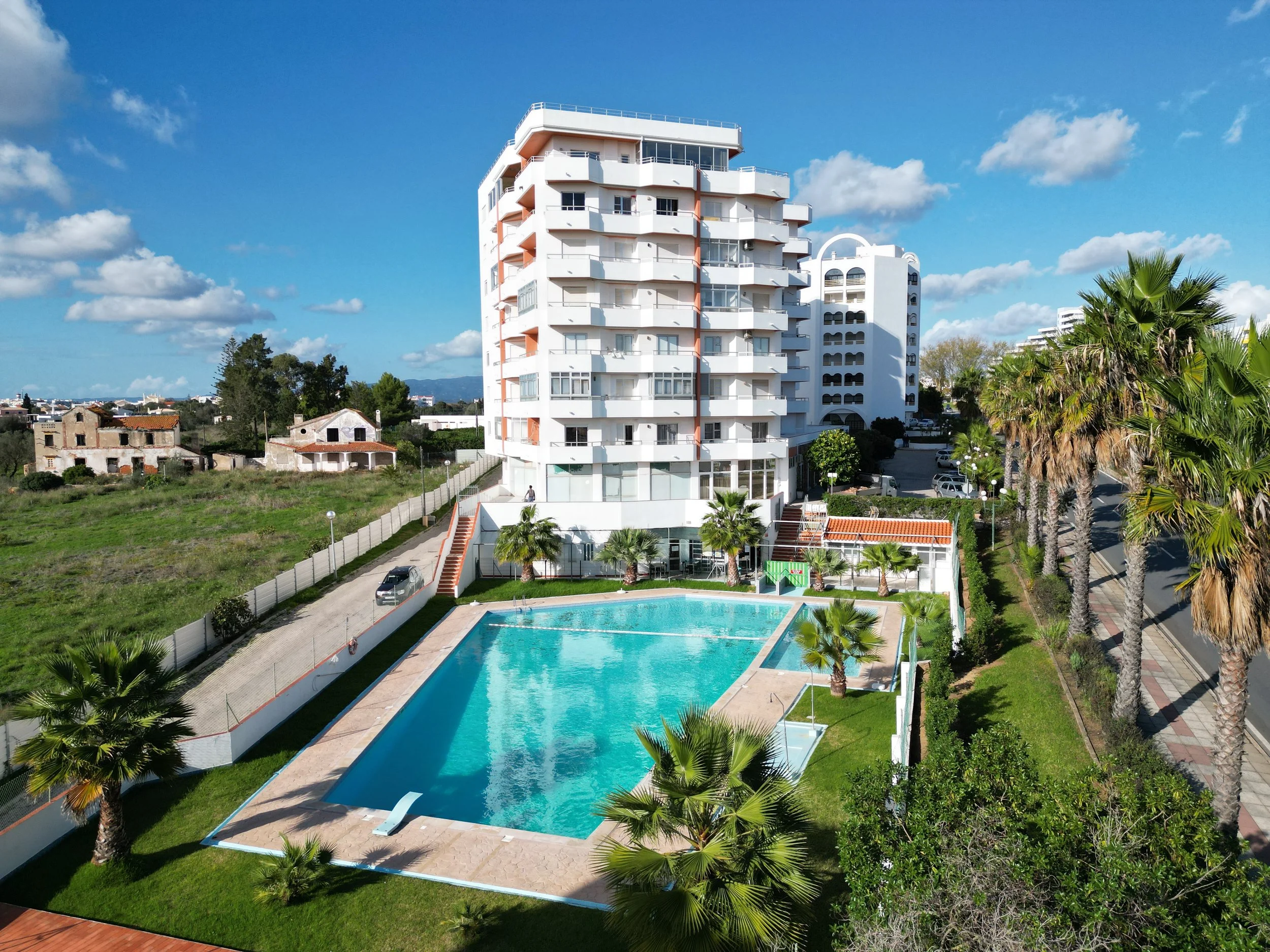 Prédio residencial com piscina ao ar livre e palmeiras ao redor, sob céu azul com algumas nuvens.