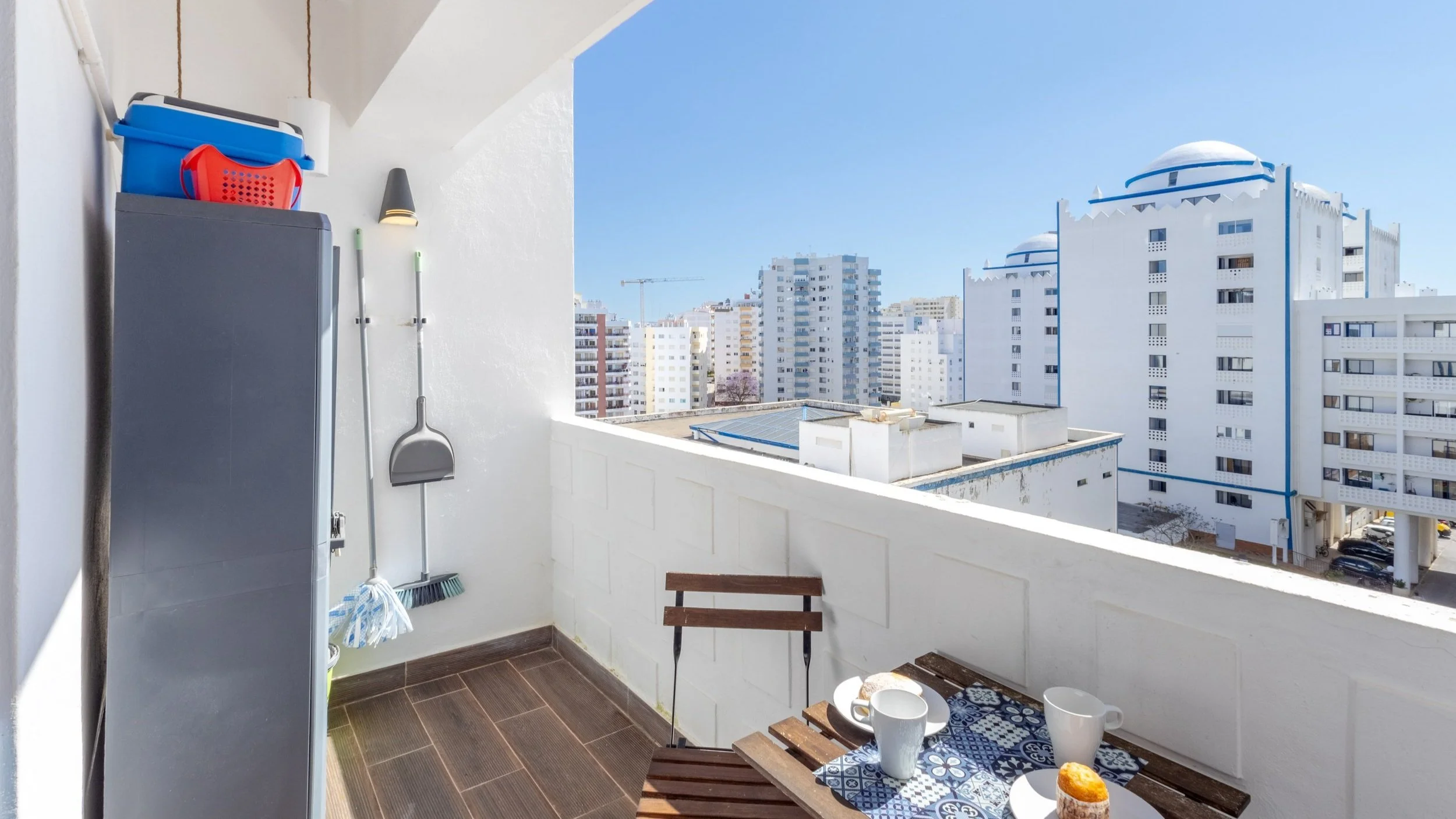 Varanda de apartamento com mesa de madeira, duas xícaras, prato com pão de queijo, cadeira de madeira, parede branca, armário com cadeira azul, balde vermelho, escova, vassoura, e vista de prédios em uma cidade ensolarada.