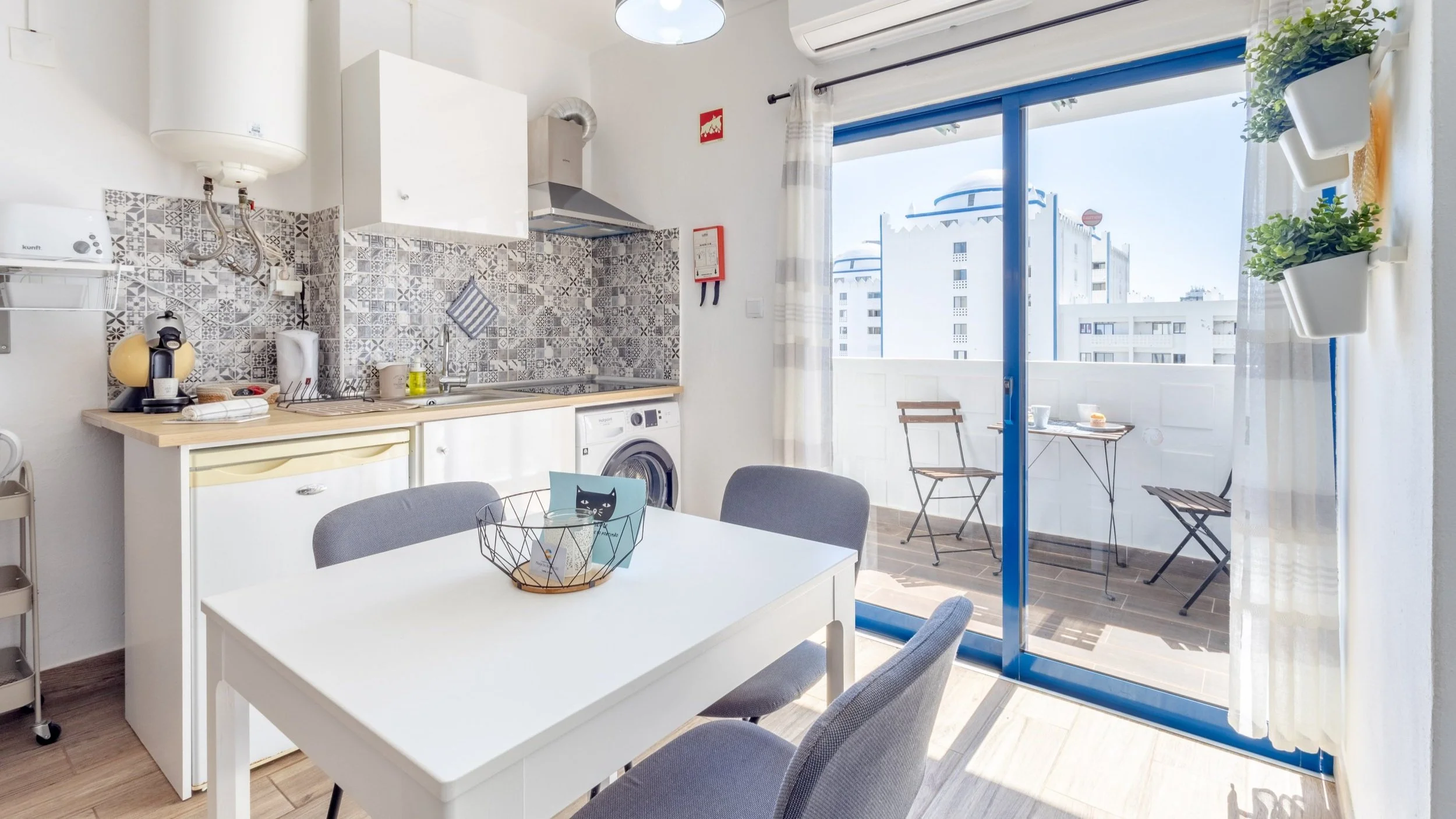 Cozinha com espaço para jantar, balcão de cozinha, balcão pequeno com máquina de café, parede decorada com azulejos em cinza, mesa branca com três cadeiras cinza, varanda com duas cadeiras de madeira e uma pequena mesa, piso de madeira, parede branca com janelas de vidro, cortinas brancas e plantas decorativas.