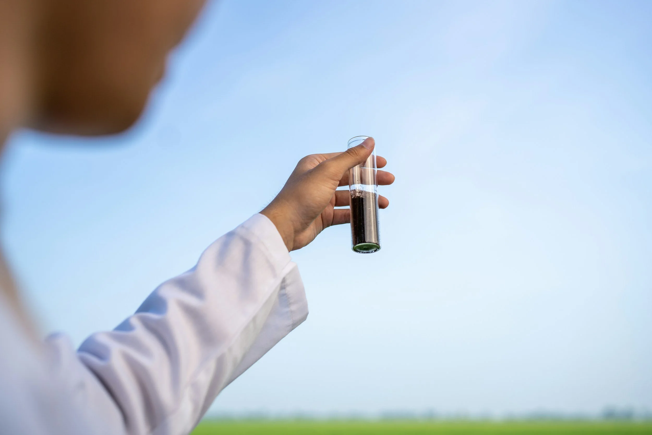 A person in a white lab coat holding a test tube filled with a dark liquid outdoors against a clear blue sky.