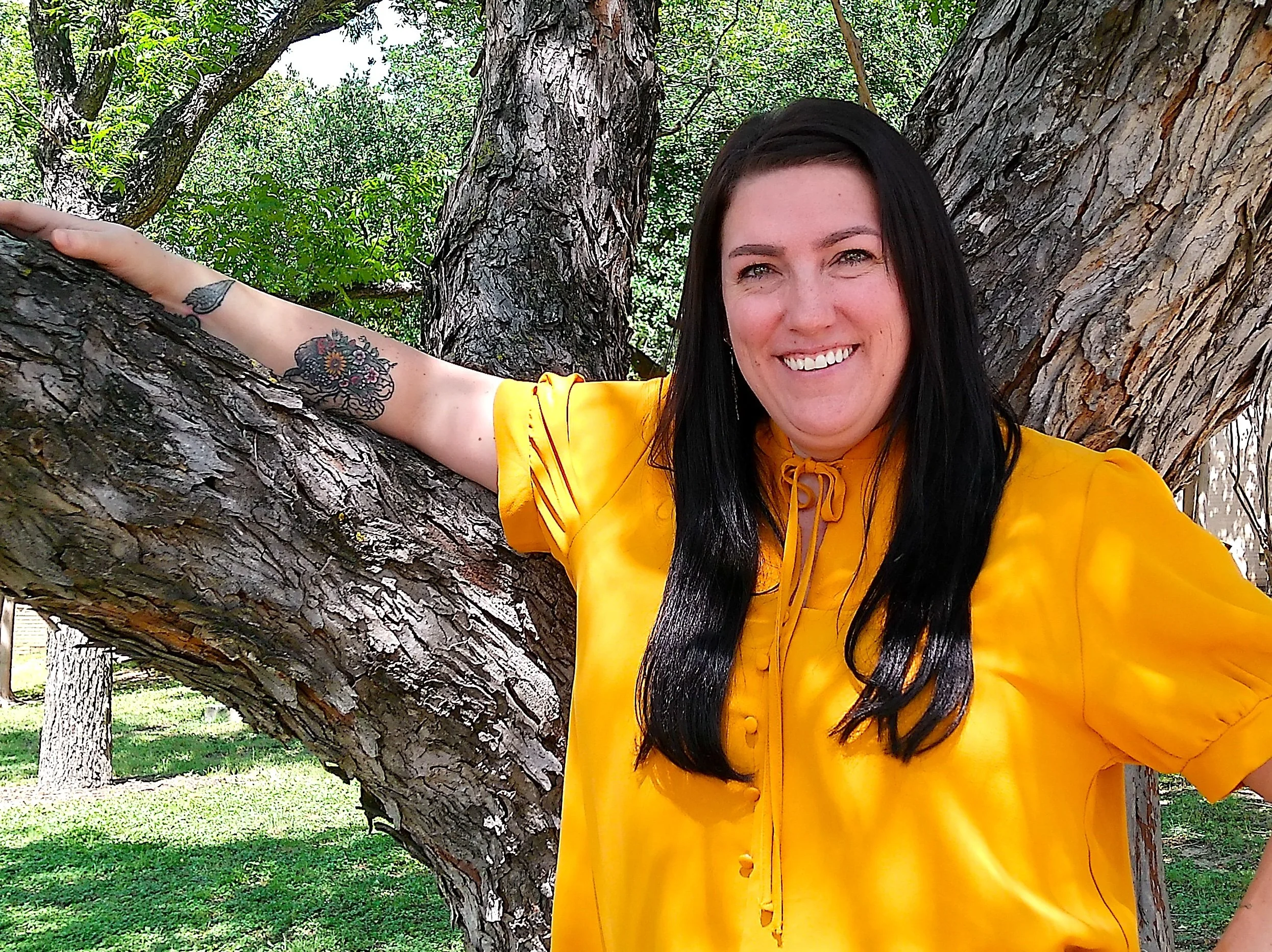 Smiling woman with black hair, wearing a yellow blouse, standing by a large tree with outstretched arms, in a park with green grass and trees in the background.