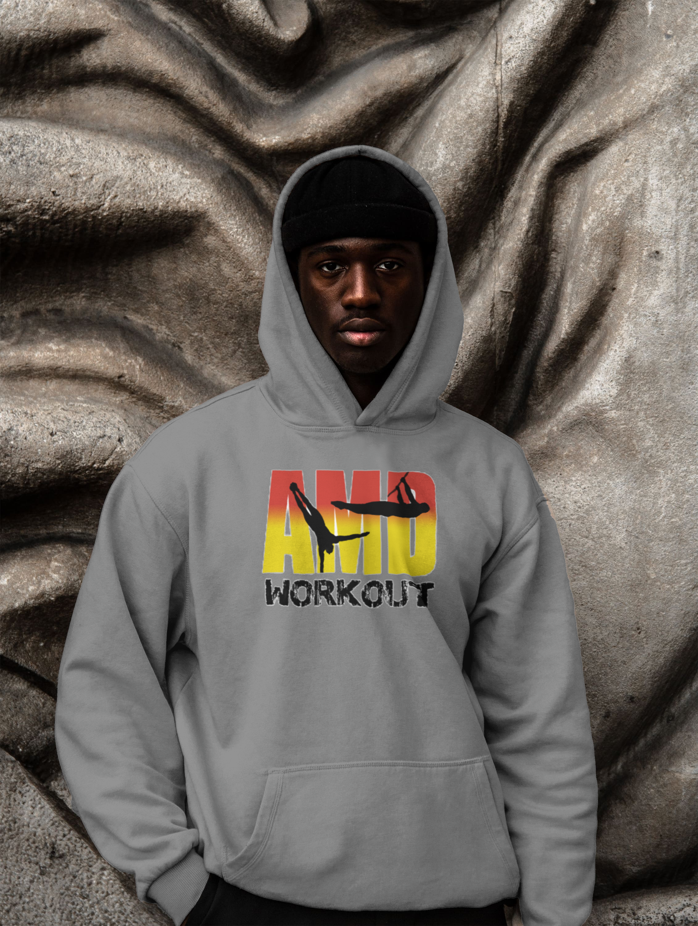 Jeune homme portant un hoodie gris avec un dessin d'acrobates et le texte 'AMP WORKOUT', posant devant une sculpture en pierre