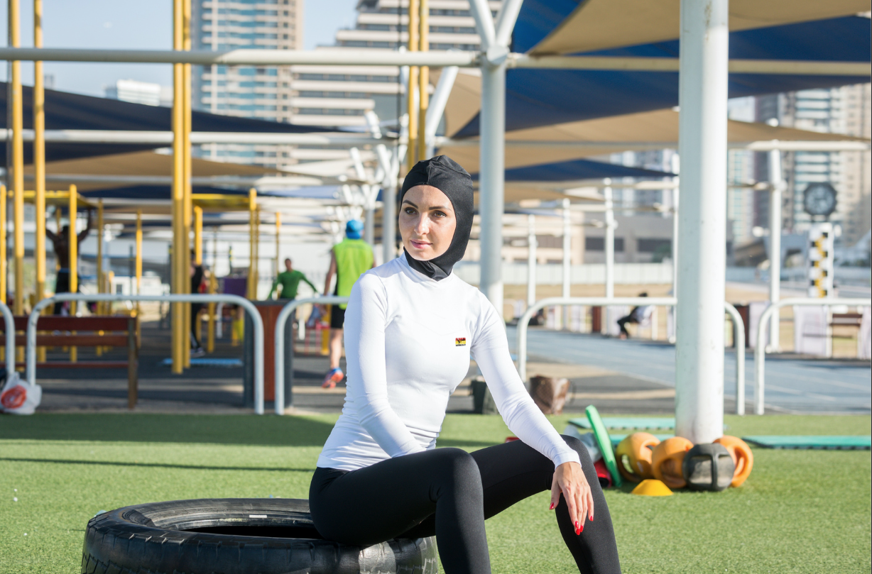 Une femme portant un casque de natation, vêtue d'une tenue de sport blanche et noire, assise sur un pneu dans une salle d'entraînement en plein air avec des équipements de fitness autour.