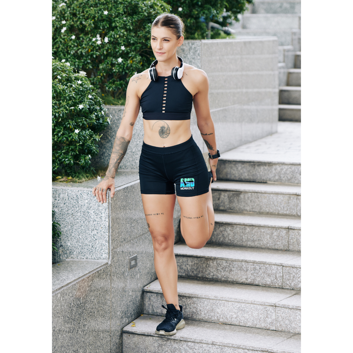 Sportive qui s'étire sur un escalier en pierre, en tenue de sport noire AMD WORKOUT, un casque audio autour du cou