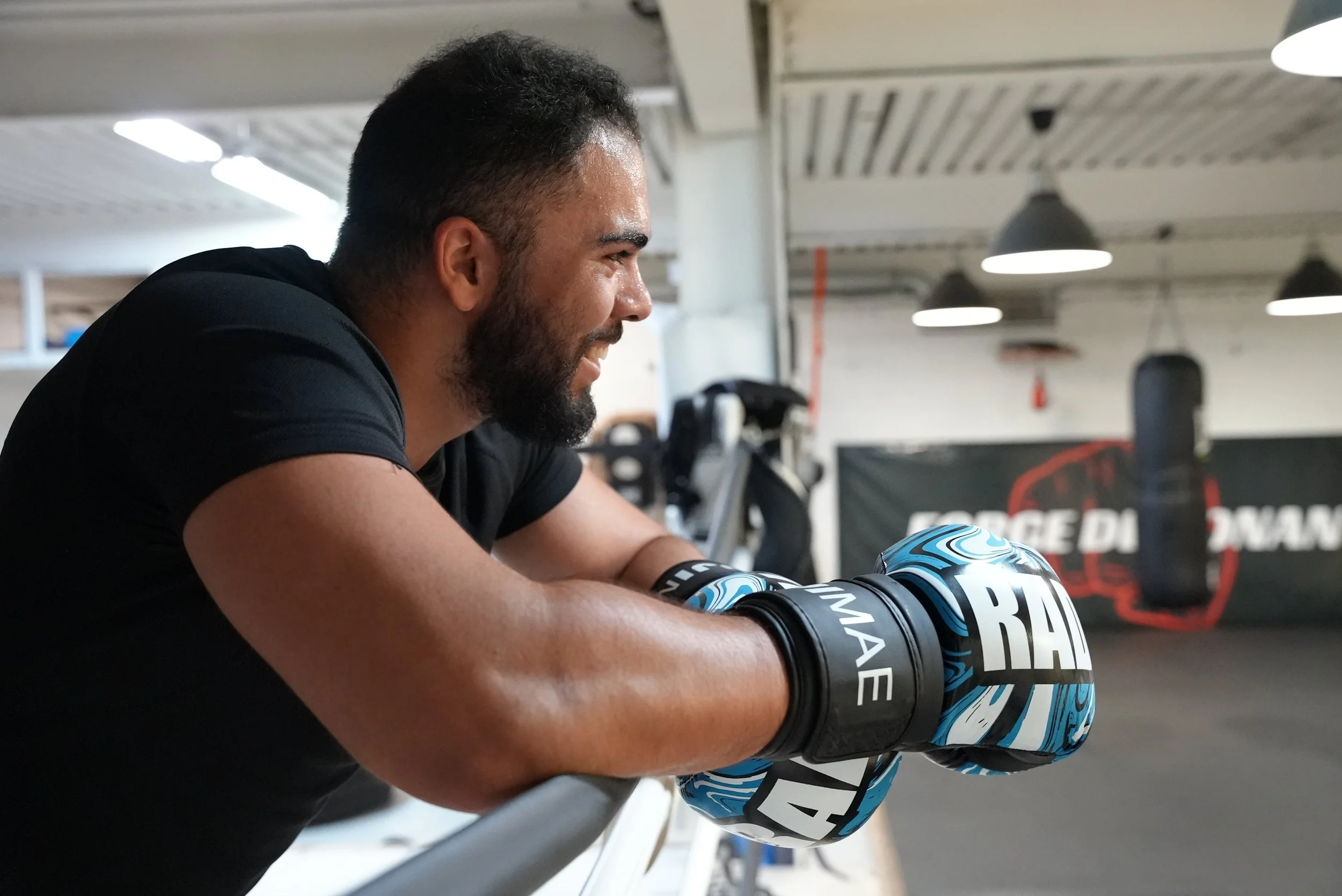 Un homme souriant à l'entraînement de boxe dans une salle de sport, avec des gants de boxe bleu et noir. Il est concentré et s'exerce, avec des équipements de boxe en arrière-plan.