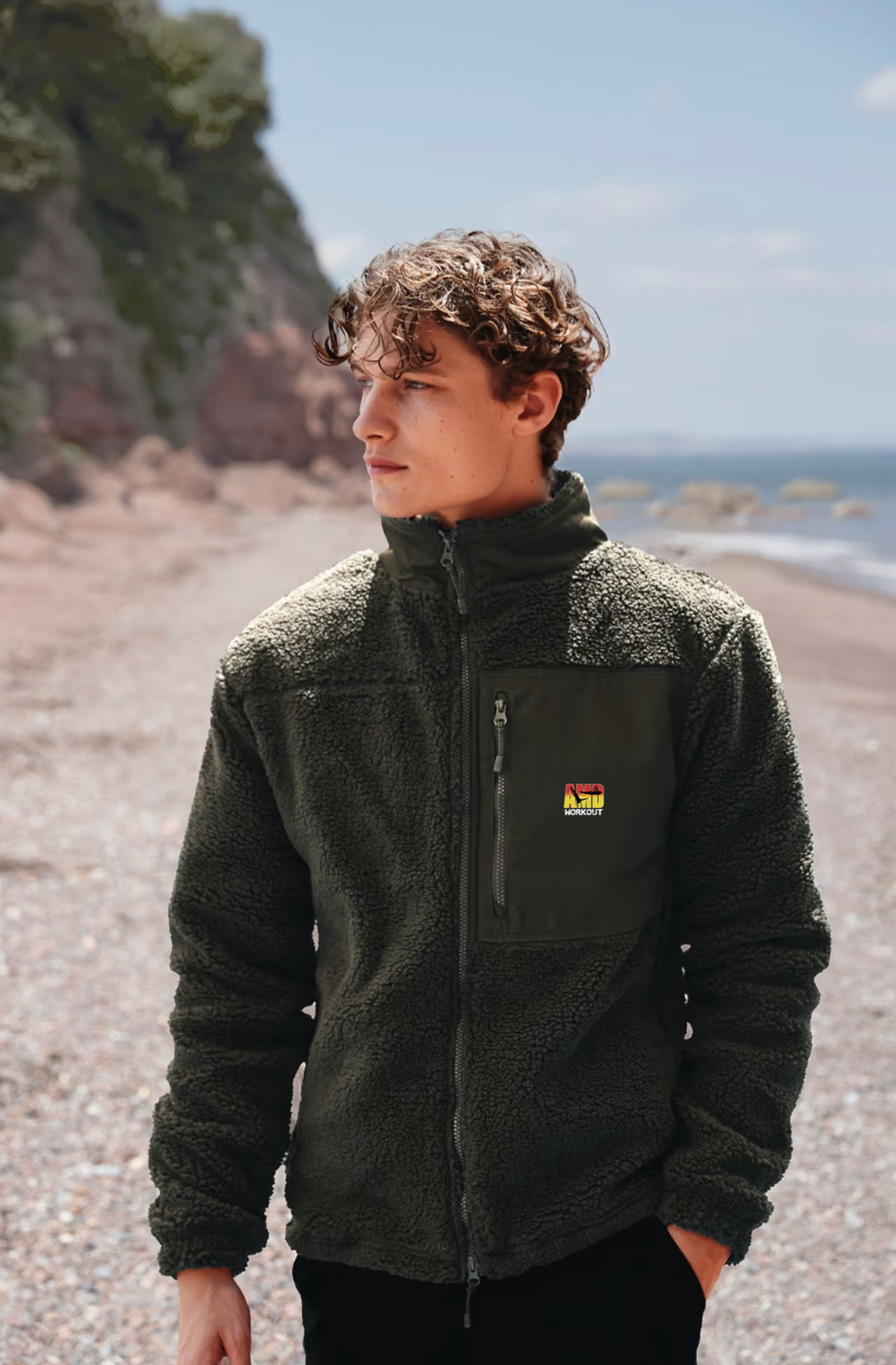 Un jeune homme avec des cheveux bouclés porte un sweat à capuche noir en tissu pelucheux sur une plage rocheuse, avec un ciel clair et la mer en arrière-plan.