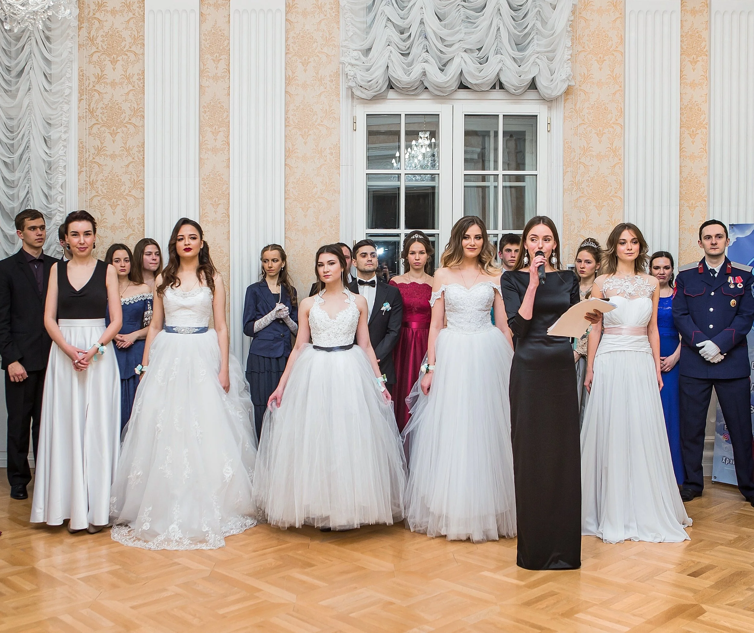 Group of young women and one young man in formal dresses and suits standing in a line indoors at a formal event, with a woman speaking into a microphone.